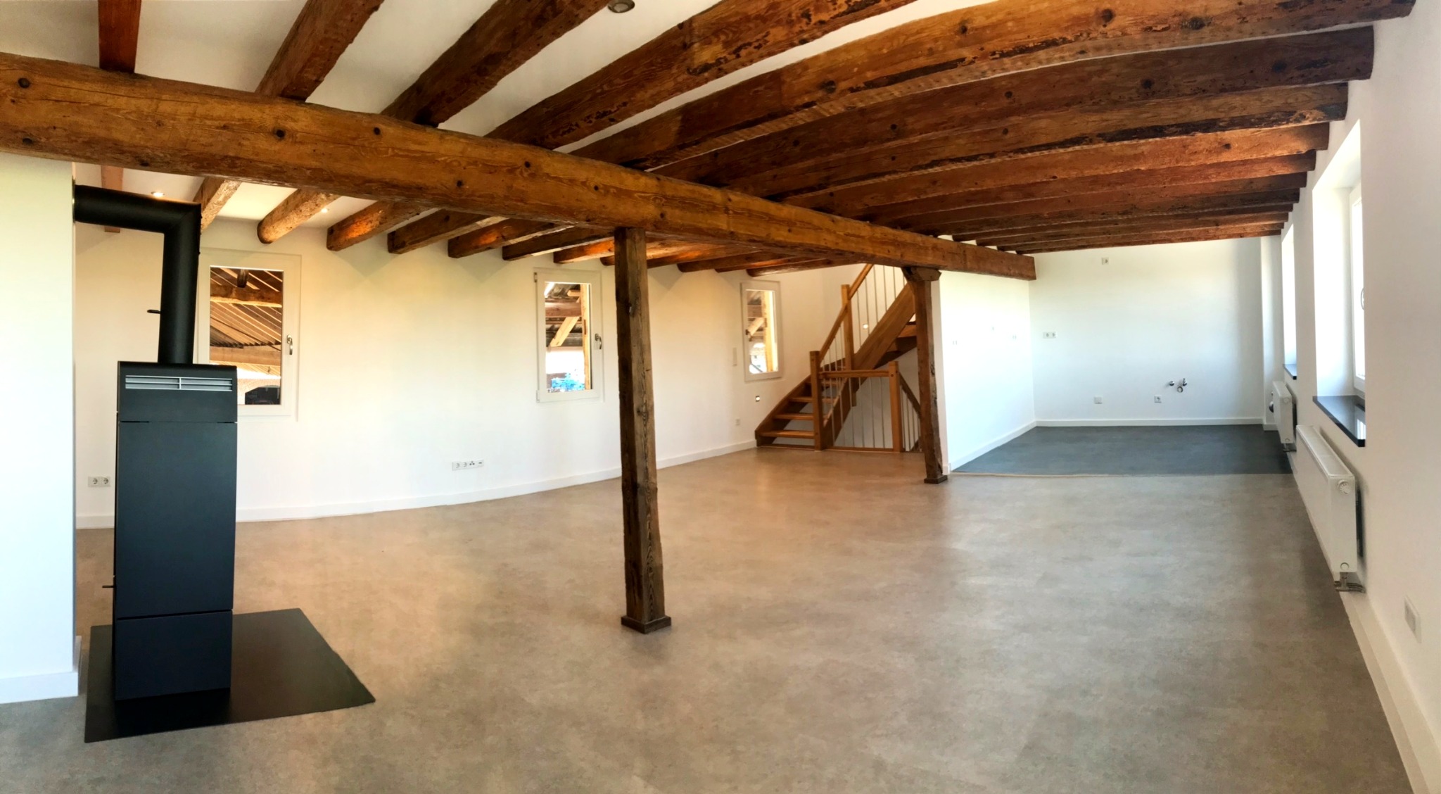 Przestronne wnętrze po remoncie z odsłoniętymi drewnianymi belkami stropowymi, nową podłogą, grzejnikami, piecem wolnostojącym i drewnianymi schodami.