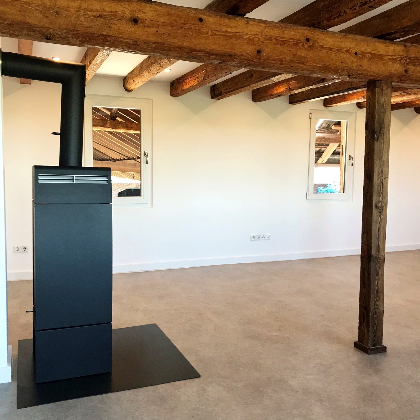 Wnętrze pokoju z nowoczesnym, czarnym piecem wolnostojącym na tle białych ścian, drewnianych belek stropowych i kolumny, widoczne gniazdka elektryczne i okna z widokiem na konstrukcję dachu.