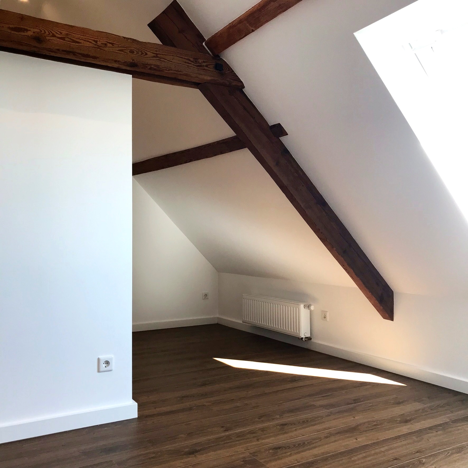 Jasne poddasze z drewnianą podłogą, białymi ścianami, grzejnikiem i widocznymi belkami stropowymi, oświetlone promieniami słońca wpadającymi przez okno dachowe.