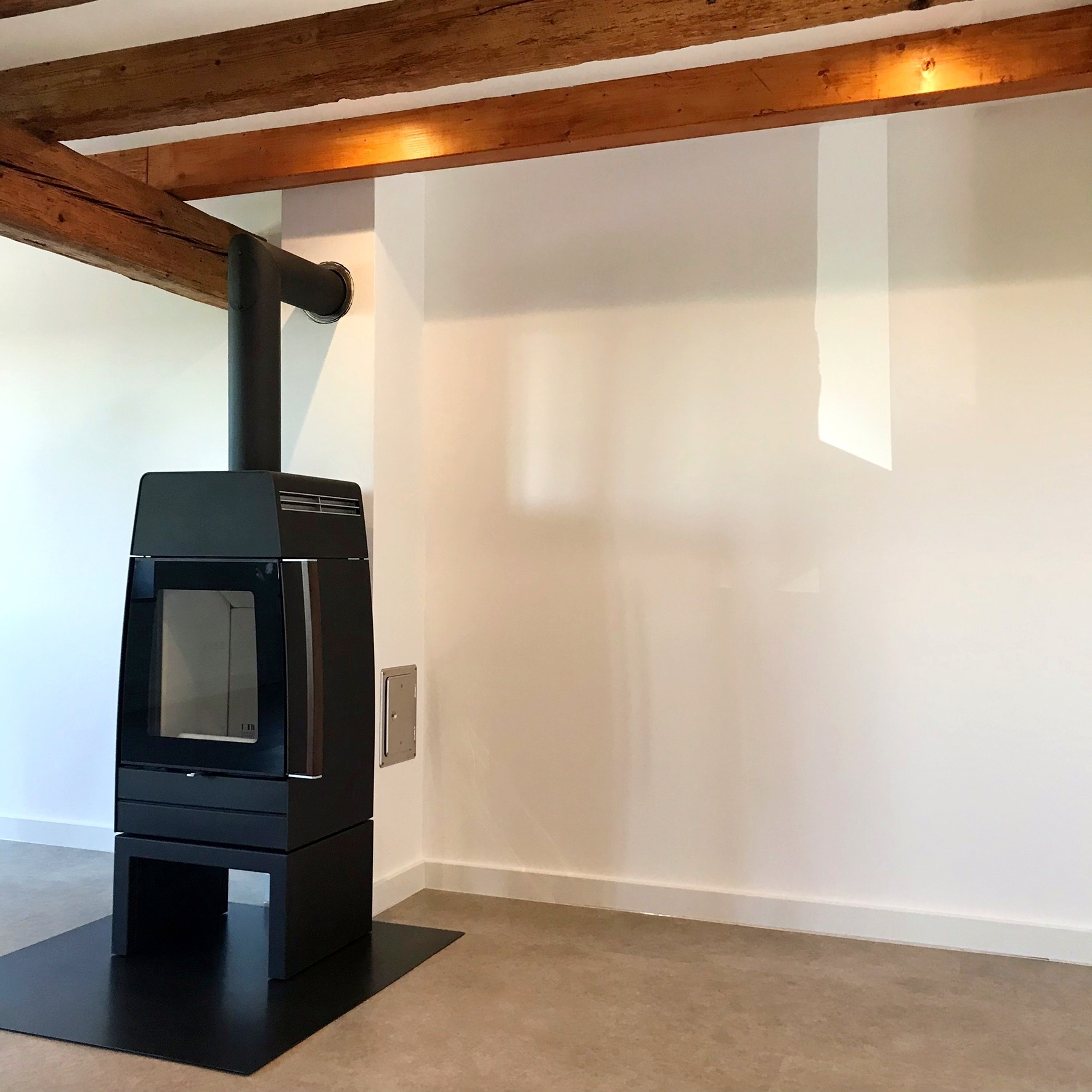 Nowoczesny czarny piec wolnostojący z czarnym kominem na tle białej ściany i drewnianych belek stropowych.