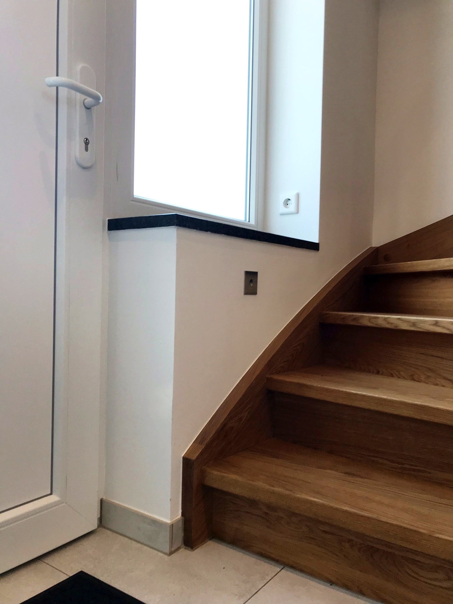 Drewniane schody z dębowymi stopniami w jasnym wnętrzu, widoczne gniazdko elektryczne na ścianie i fragment białych drzwi.