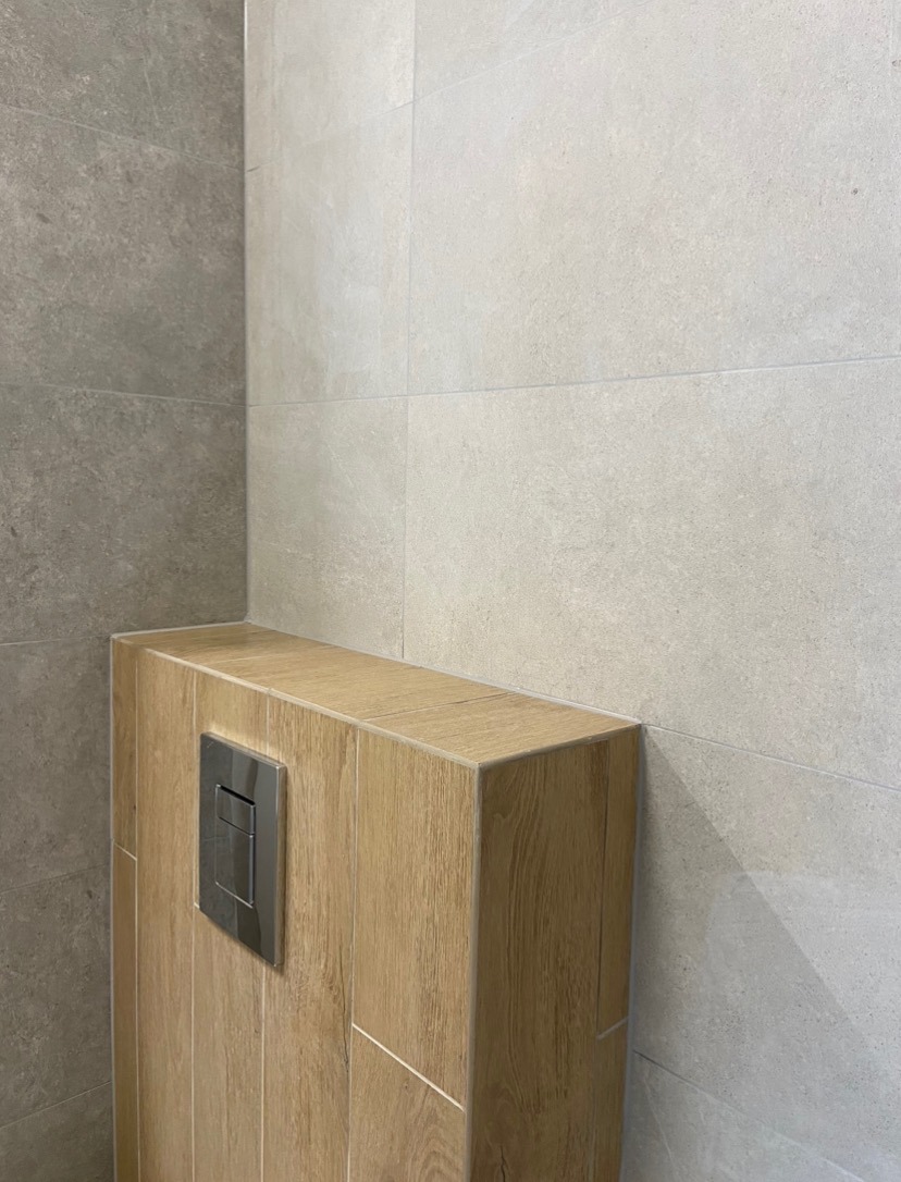 Szare płytki ścienne w łazience z zabudową geberitu wykończoną płytkami imitującymi drewno i chromowanym przyciskiem spłukującym.