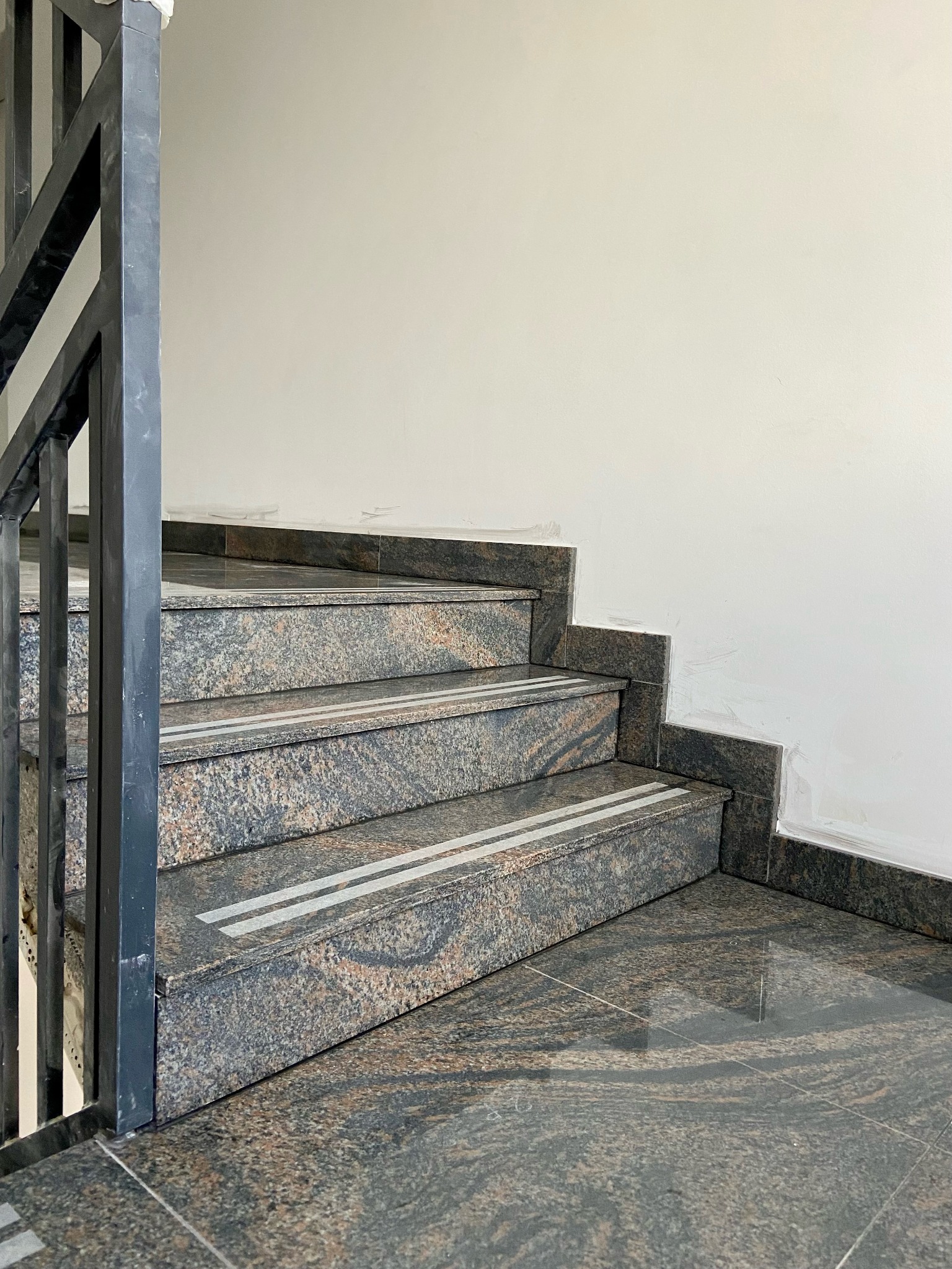 Granitowe schody z metalową balustradą, wykończone białą listwą przy ścianie, z antypoślizgowymi paskami na stopniach.