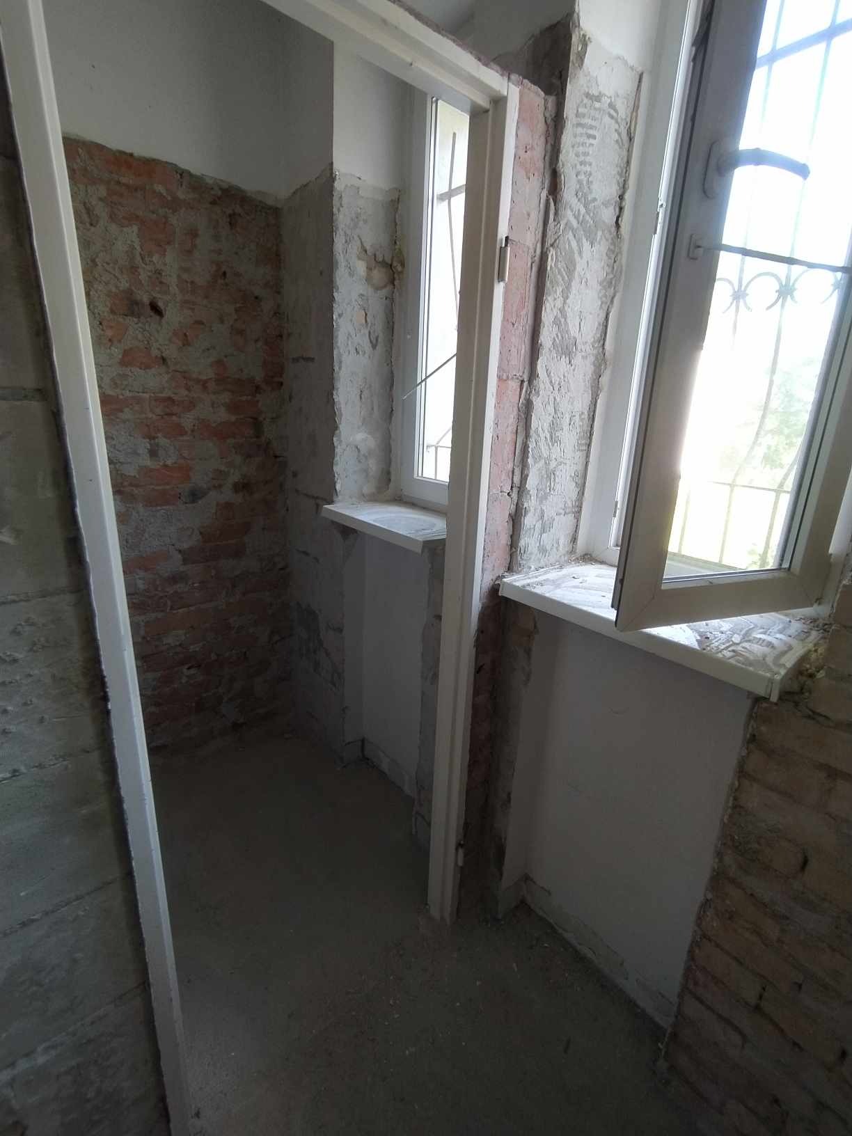 Wnętrze pomieszczenia w trakcie prac remontowych, widoczne odsłonięte cegły, surowy tynk, nowe okna z białymi ramami i parapetami, oraz ościeżnice drzwiowe przed wykończeniem.