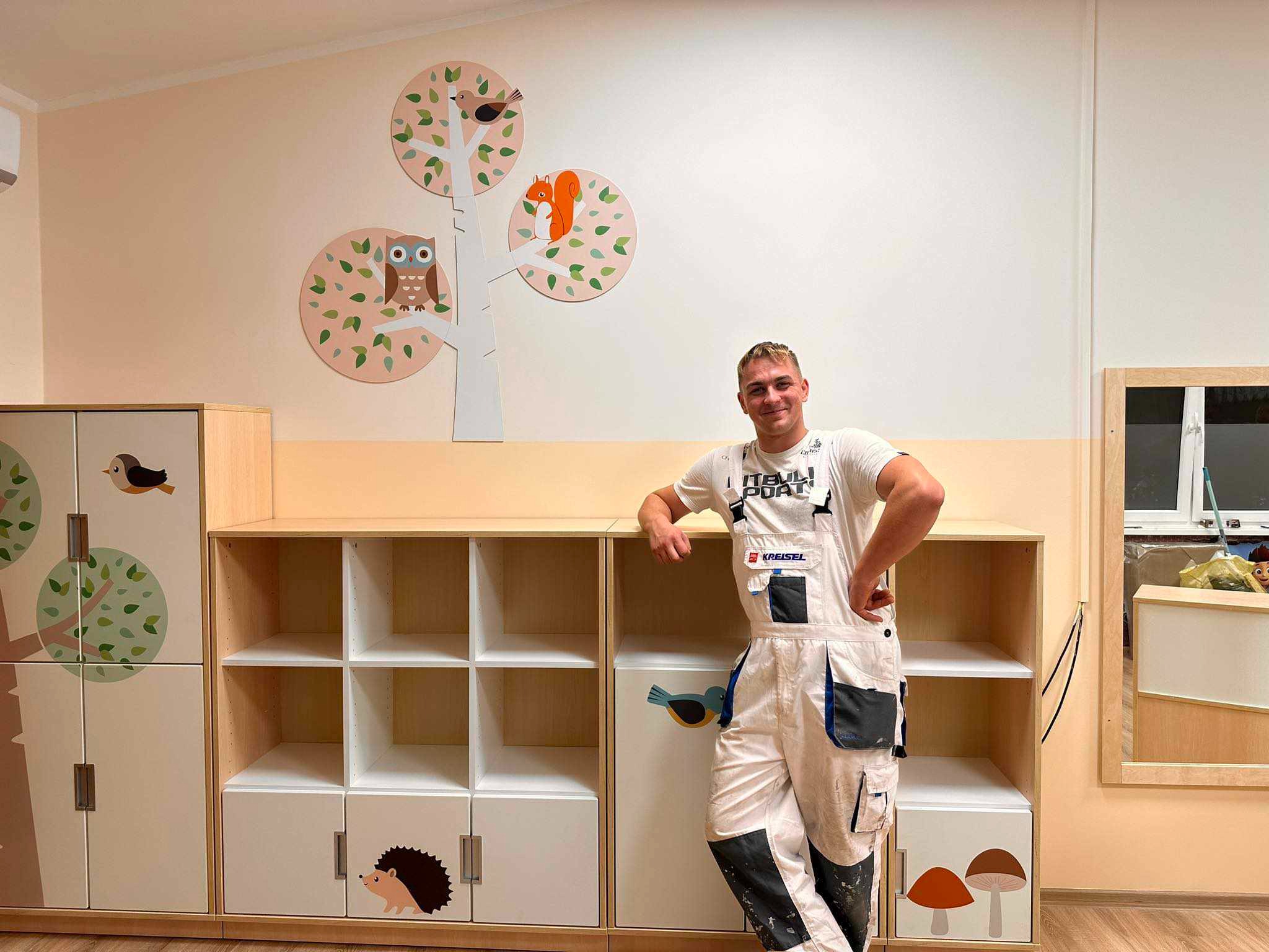 Uśmiechnięty malarz w ubraniu roboczym, oparty o regał z motywami zwierząt, na ścianie malowidło drzewa z sową, wiewiórką i ptakami, pokój dziecięcy po malowaniu.