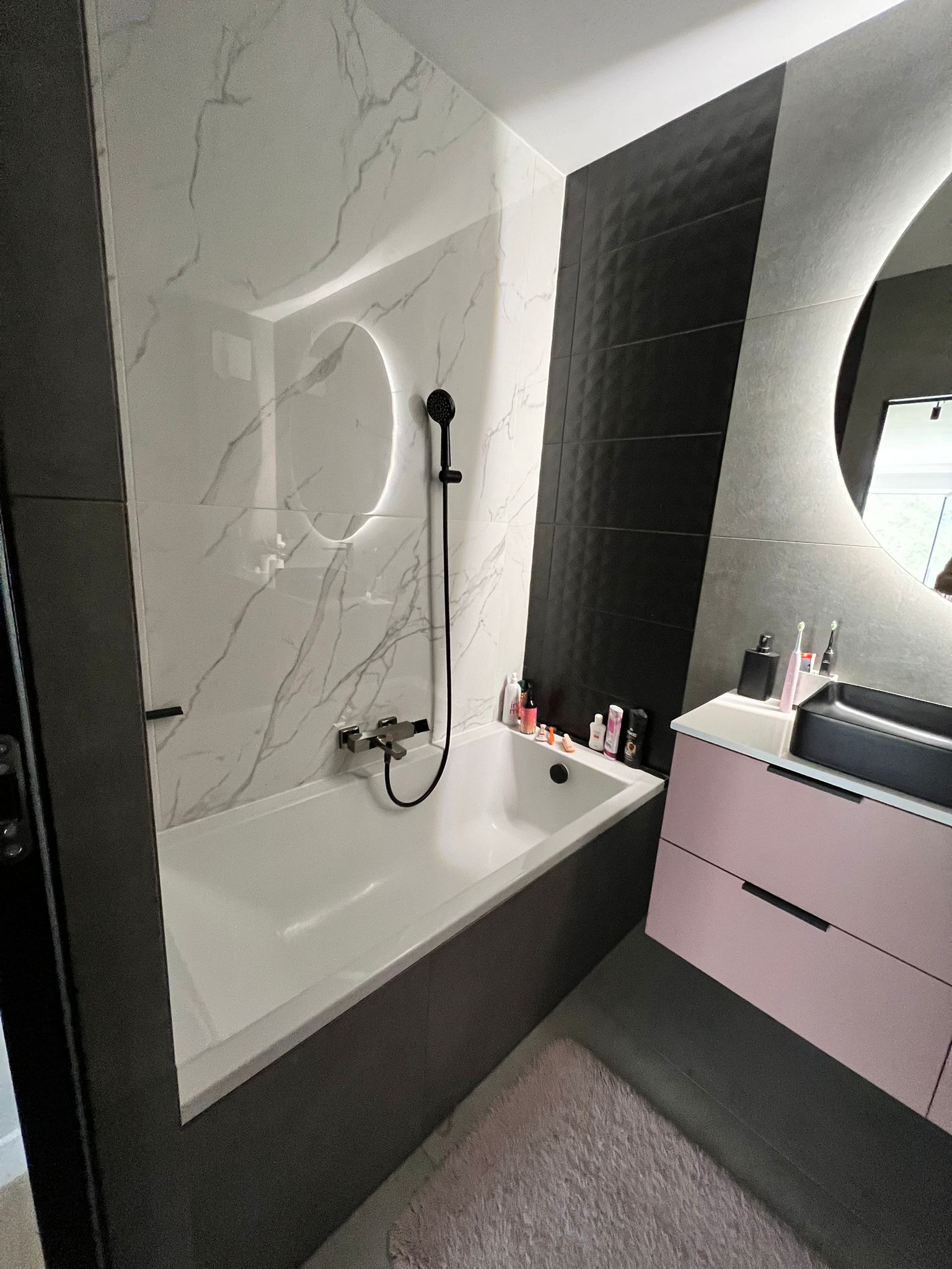 Nowoczesna łazienka z wanną obudowaną ciemnoszarymi płytkami, białe marmurowe płytki na ścianie, czarna armatura, umywalka wpuszczana w blat z szafką w kolorze pudrowego różu, okrągłe lustro...