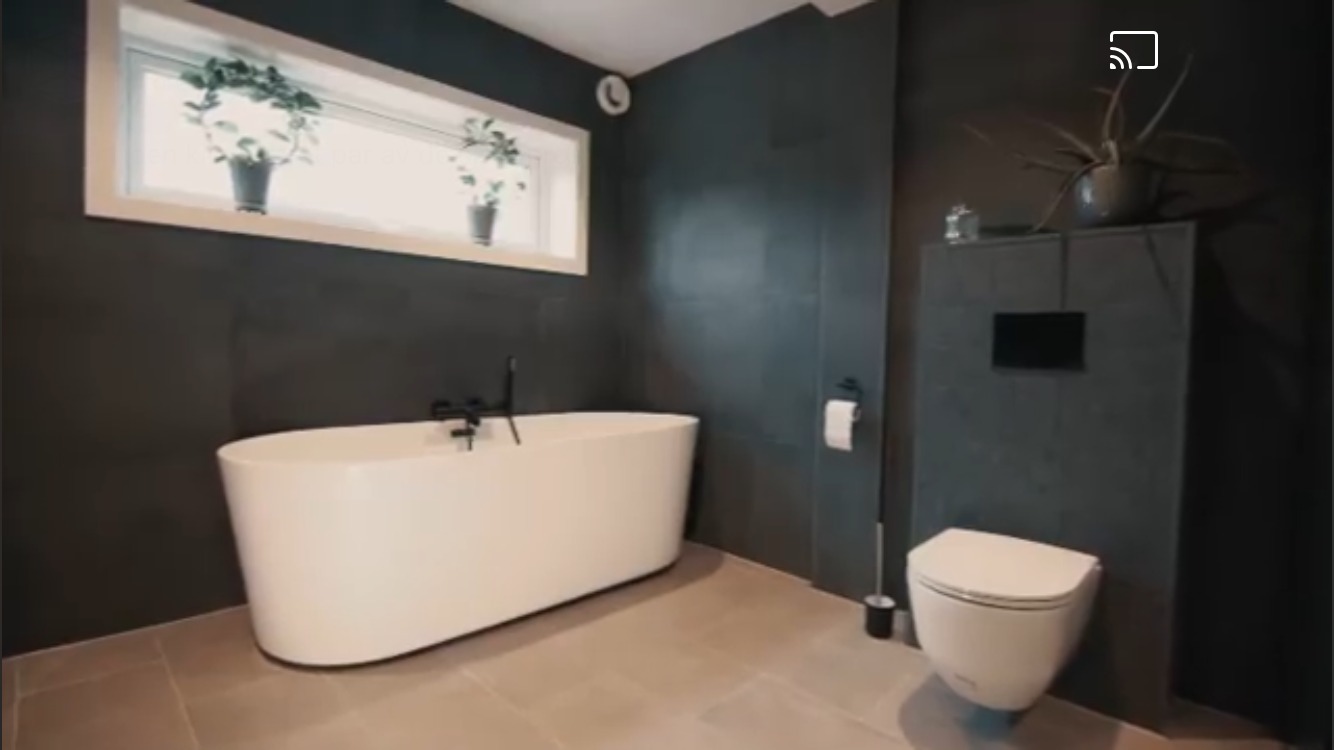 Nowoczesna łazienka z wolnostojącą białą wanną, czarną armaturą, szarymi płytkami na ścianach i podłodze, oraz roślinami w doniczkach na parapecie okna.