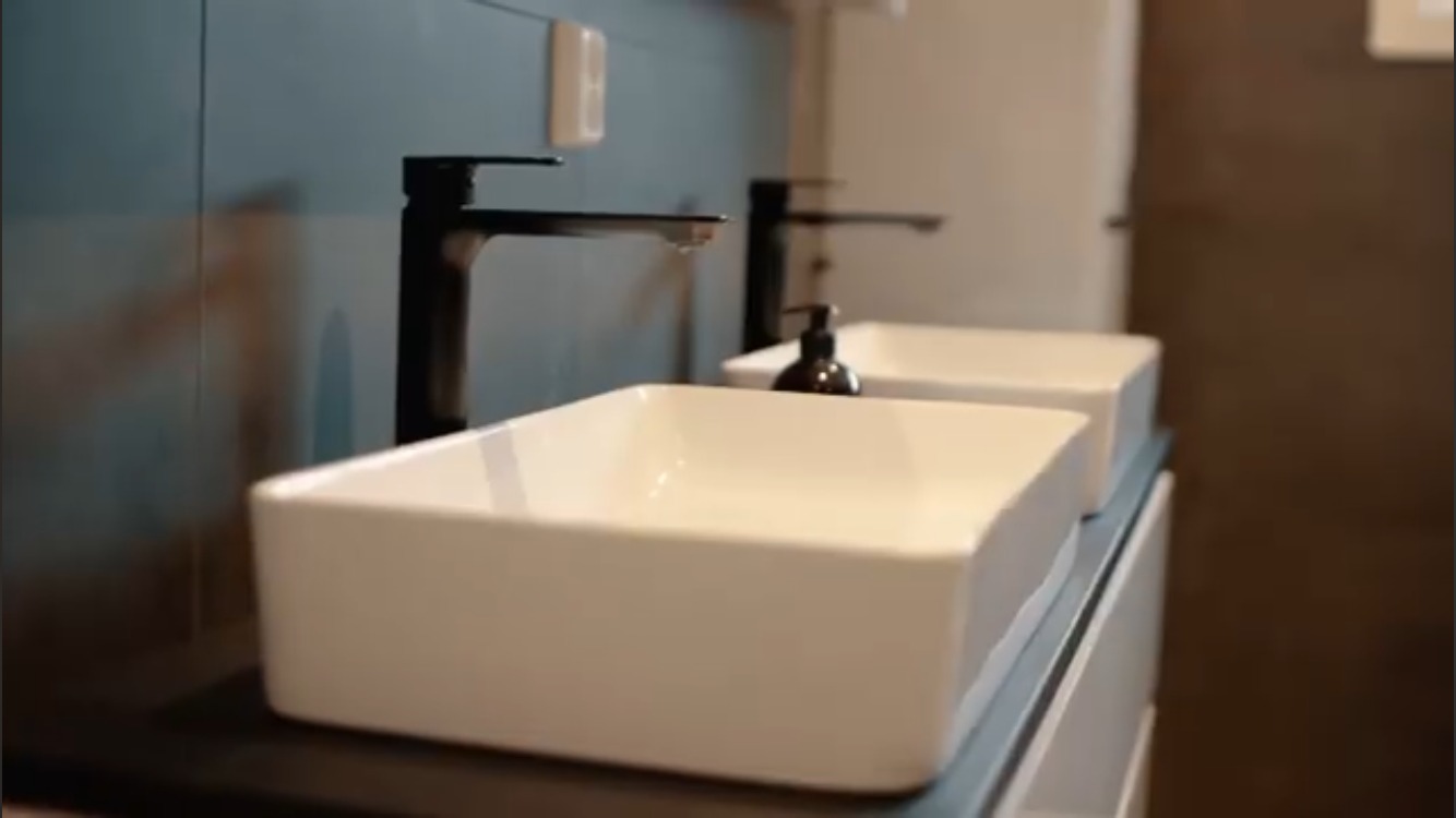 Dwie białe, prostokątne umywalki nablatowe z czarnymi, minimalistycznymi bateriami w nowoczesnej łazience z niebieskimi płytkami i brązową ścianą w tle. Z jednej baterii kapie woda.