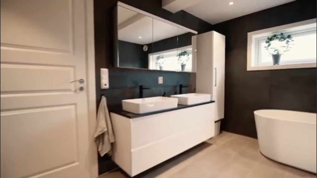 Nowoczesna łazienka z dwoma białymi umywalkami na szafce, czarnymi bateriami, lustrem z szafką, białą wanną wolnostojącą i oknem z rośliną doniczkową, wykończona w ciemnych kolorach.