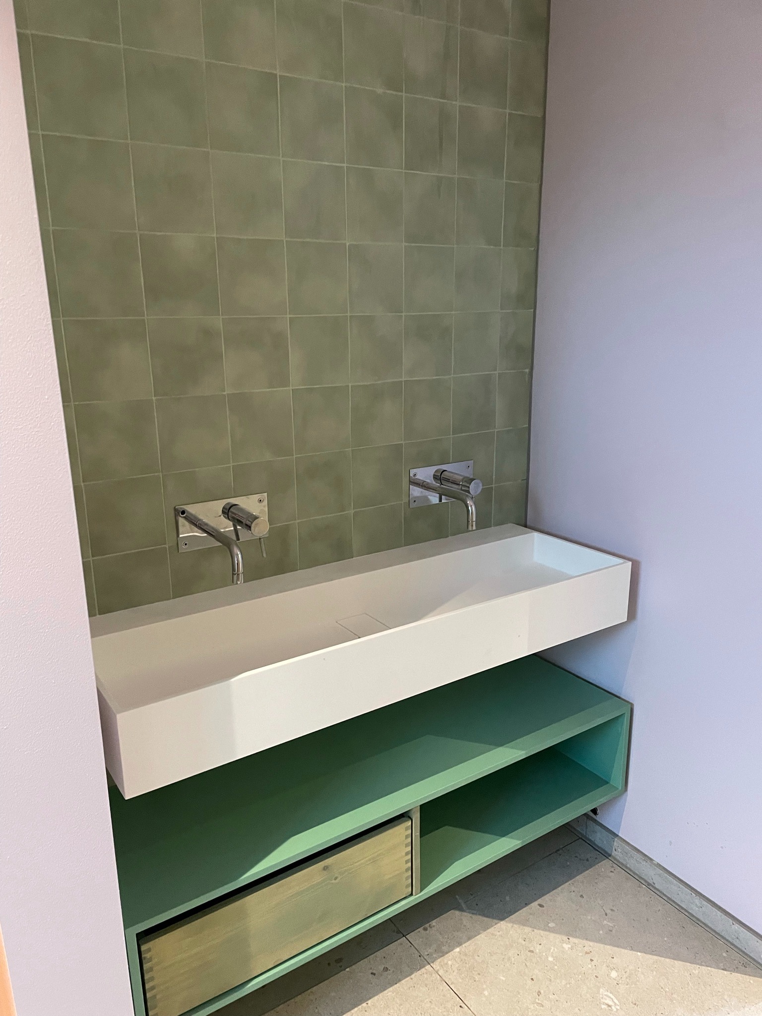 Nowoczesna łazienka z zielonymi kwadratowymi płytkami na ścianie, białą umywalką nablatową z dwoma chromowanymi bateriami, zieloną szafką pod umywalką i fioletowymi ścianami.