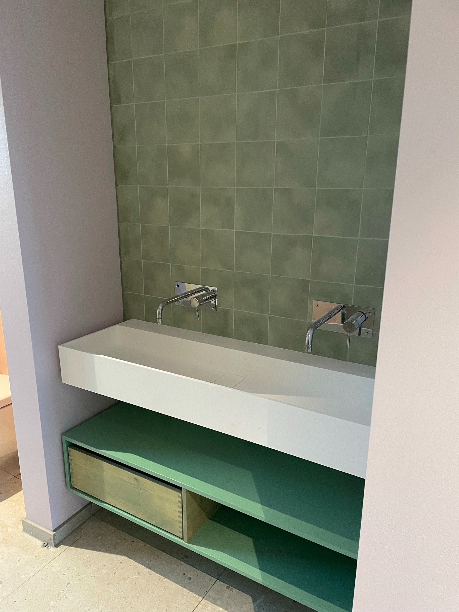 Nowoczesna łazienka z zielonymi płytkami i minimalistyczną białą umywalką nablatową z dwoma chromowanymi bateriami ściennymi, szafka pod umywalką z szufladą w odcieniach zieleni.