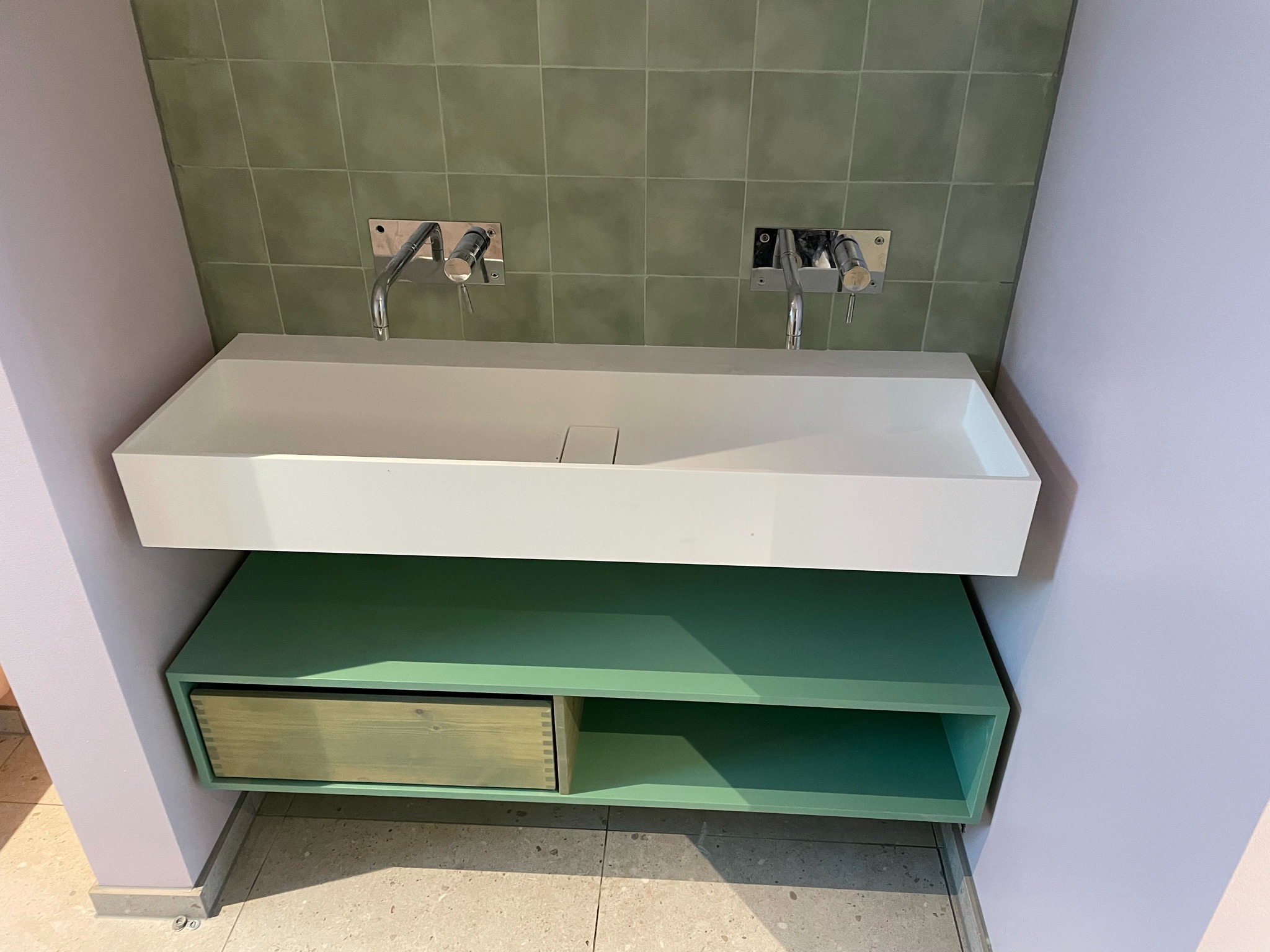Nowoczesna łazienka z białą, prostokątną umywalką nablatową, zamontowaną na zielonej szafce z szufladą z surowego drewna. Na ścianie zielone, kwadratowe płytki i chromowane baterie ścienne.