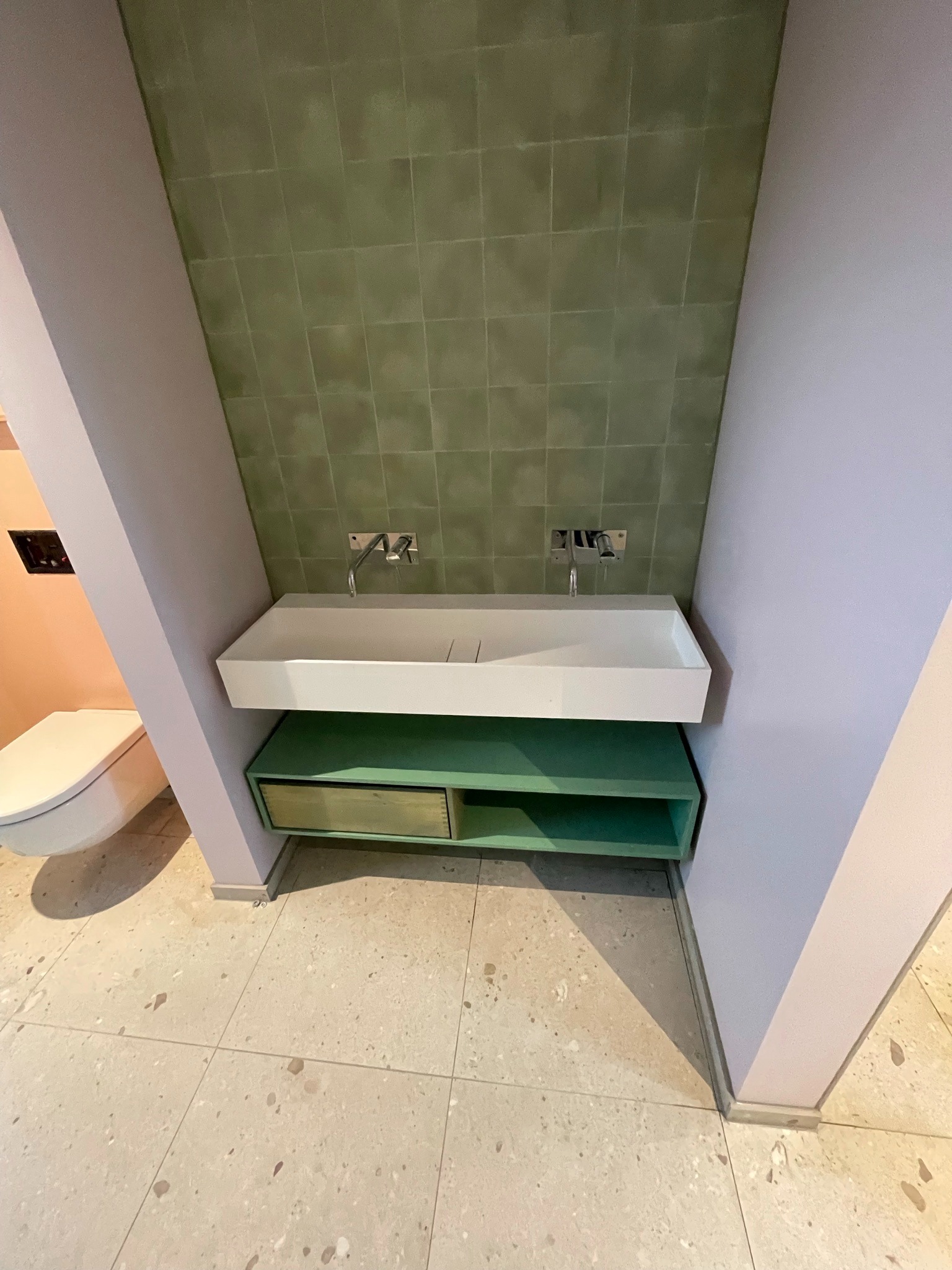 Nowoczesna łazienka z podwójną umywalką nablatową, zieloną szafką podumywalkową, oliwkowymi płytkami na ścianie oraz terazzo na podłodze, całość utrzymana w pastelowej kolorystyce.