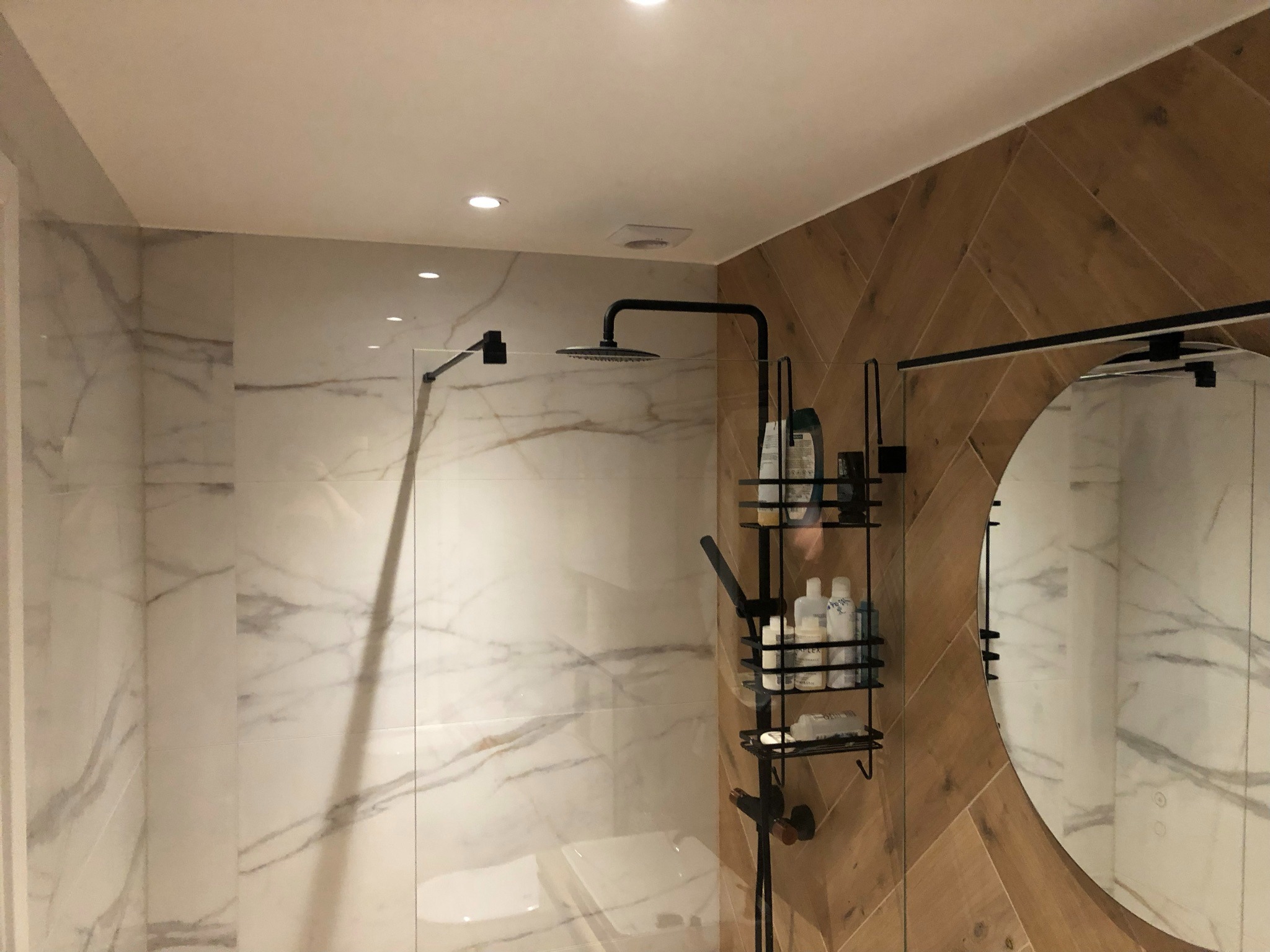 Nowoczesna łazienka z marmurowymi płytkami na ścianach, drewnopodobnymi płytkami ułożonymi w jodełkę, okrągłym lustrem i czarną armaturą prysznicową z półkami na kosmetyki.