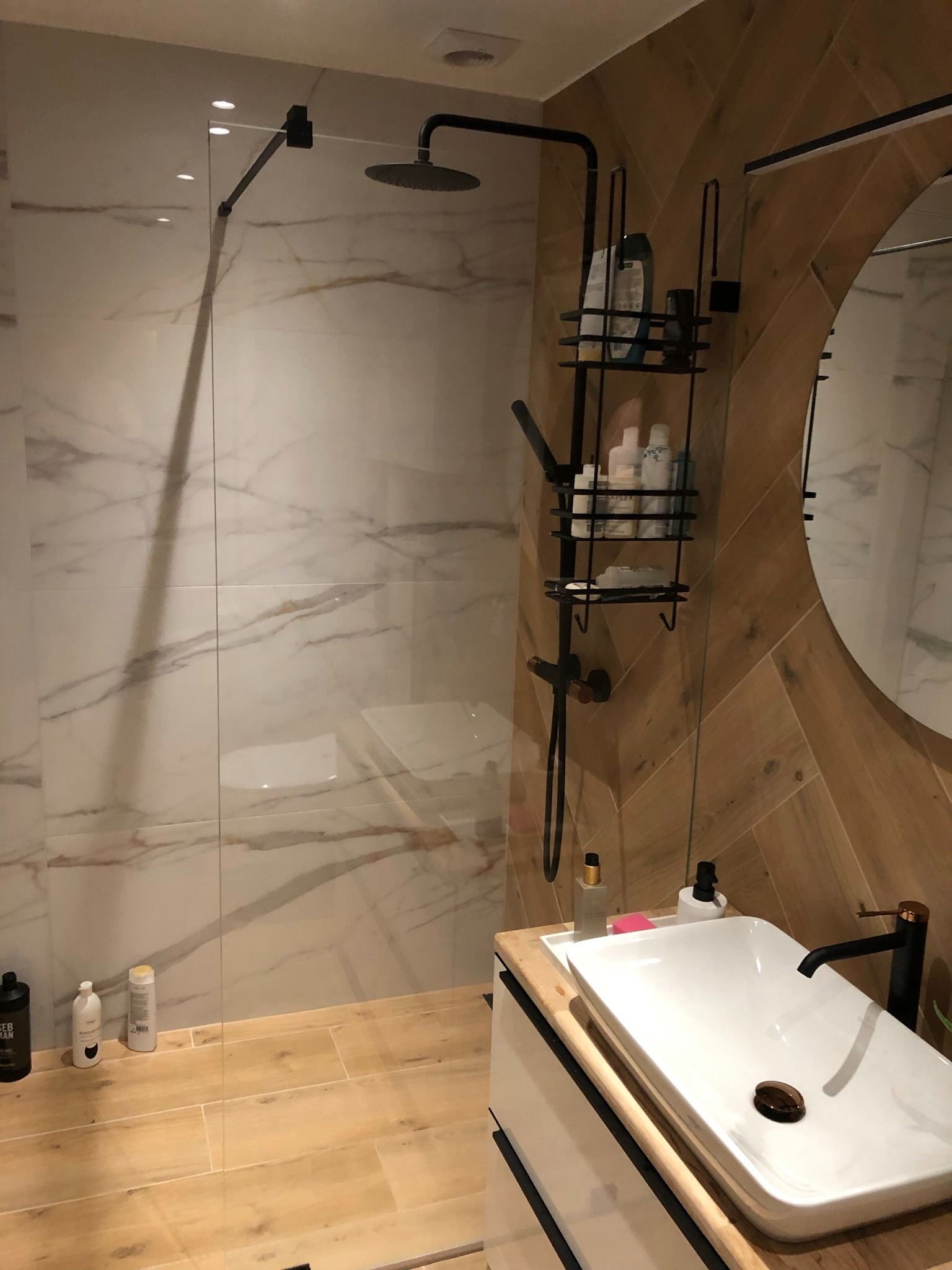 Nowoczesna łazienka w Rumi z kabiną prysznicową typu walk-in, wykończona płytkami imitującymi marmur i drewno, z czarną armaturą i minimalistyczną umywalką nablatową.