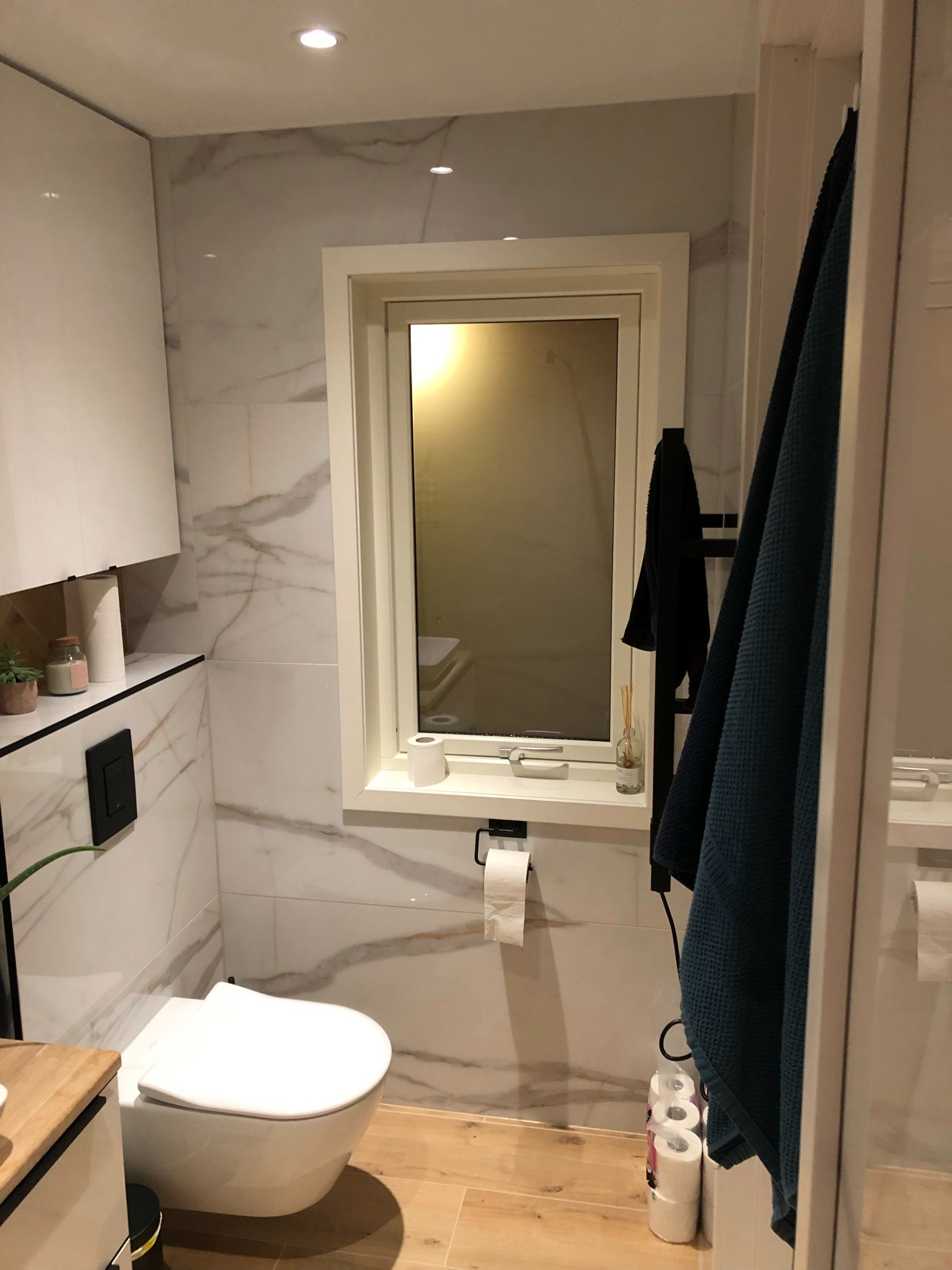 Nowoczesna łazienka z wiszącą toaletą, marmurowe płytki na ścianach, drewnopodobna podłoga, okno z białą ramą i dekoracyjnymi akcesoriami.