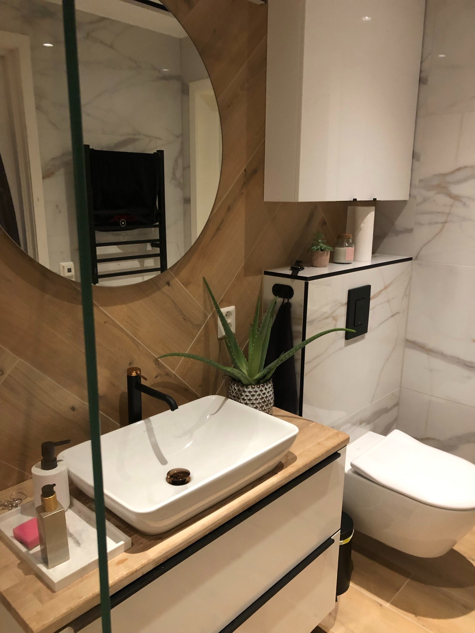 Nowoczesna łazienka z umywalką nablatową, baterią w kolorze czarnym i lustrem w okrągłej ramie z drewnopodobnych płytek ułożonych w jodełkę; w tle widoczny grzejnik drabinkowy.