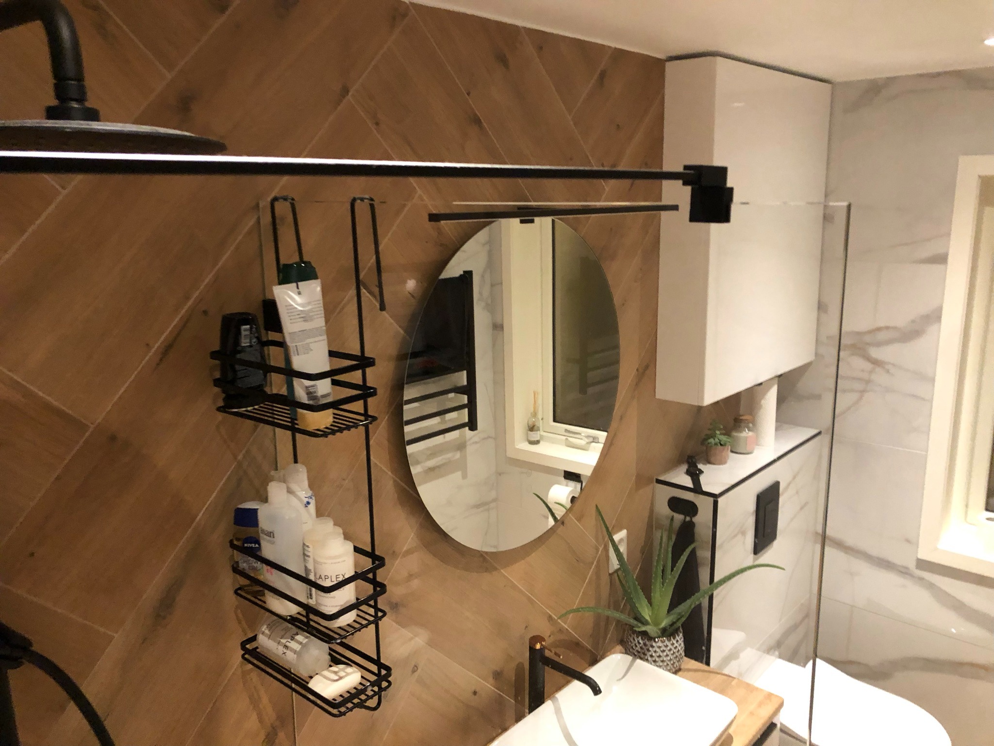 Nowoczesna łazienka z drewnopodobnymi płytkami ułożonymi w jodełkę na ścianie, czarną armaturą, okrągłym lustrem i półką na kosmetyki, widoczne również białe szafki i toaleta.