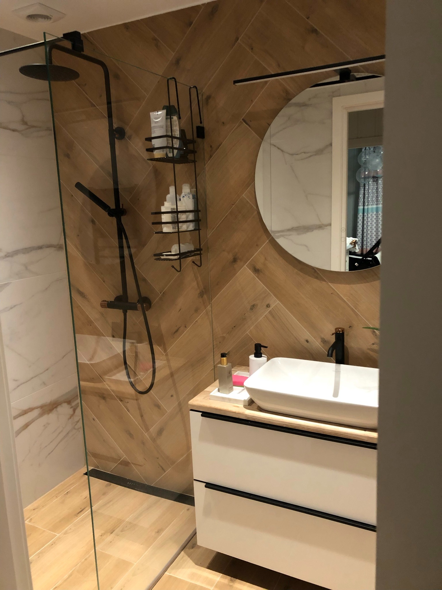 Nowoczesna łazienka z kabiną prysznicową typu walk-in, drewnopodobnymi płytkami ułożonymi w jodełkę na ścianie, okrągłym lustrem z podświetleniem LED i białą umywalką nablatową na szafce z szufladami.