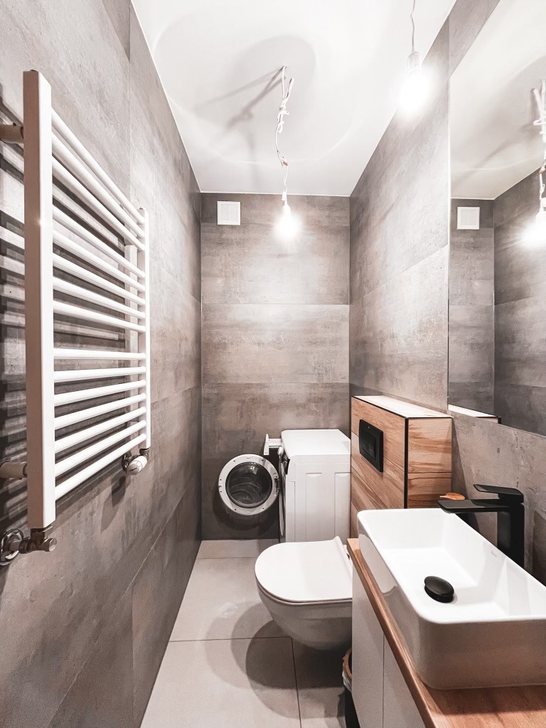Wąska łazienka z szarymi płytkami imitującymi beton, z wiszącą toaletą, pralką z suszarką, grzejnikiem drabinkowym i umywalką nablatową z czarną armaturą.
