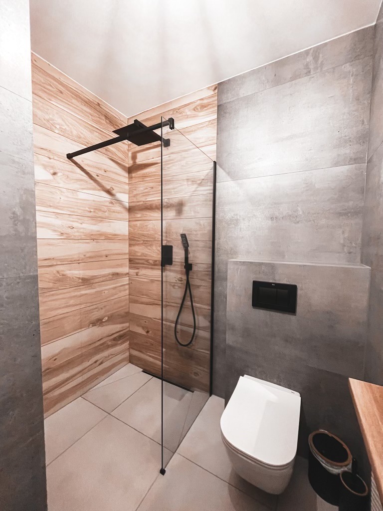 Nowoczesna łazienka z kabiną prysznicową typu walk-in z czarną armaturą, drewnopodobnymi płytkami na ścianie i szarą betonową ścianą z podtynkową spłuczką WC.