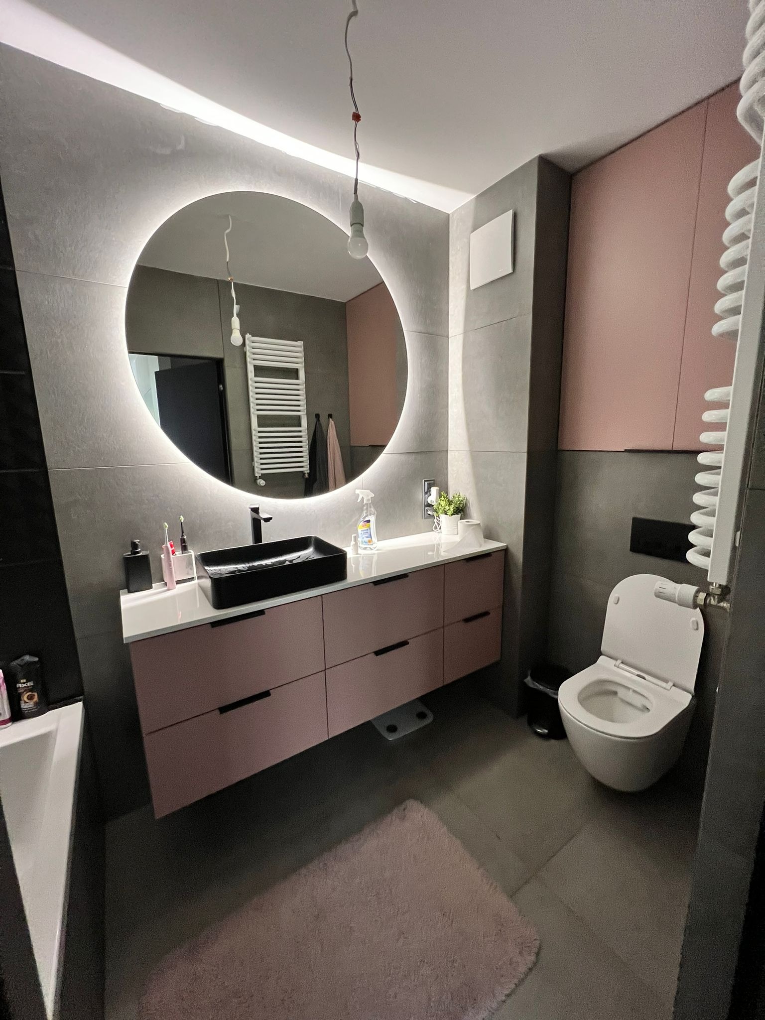 Nowoczesna łazienka w szaro-różowej kolorystyce z okrągłym lustrem podświetlanym LED, czarną umywalką nablatową i wiszącą toaletą.