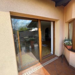 OMEGA Krzysztof Byckiewicz - Przesuwne drzwi balkonowe z brązową ramą, widok z zewnątrz na taras z terakotową podłogą i roślinami w doniczce na parapecie.