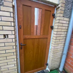 Brązowe drzwi wejściowe z szybą zamontowane w murze z cegły, widoczne ślady pianki montażowej, z zewnątrz widoczna instalacja elektryczna oraz rura spustowa.