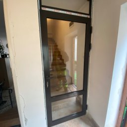 OMEGA Krzysztof Byckiewicz - Czarna aluminiowa rama drzwiowa z dużym przeszkleniem wewnętrznym, widoczna osoba w odblaskowej kamizelce na schodach w tle, ślady pianki montażowej i uszkodzenia ściany.