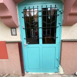 OMEGA Krzysztof Byckiewicz - Turkusowe drzwi wejściowe z czarną kratą, osadzone w różowej fasadzie budynku, widoczne ślady pianki montażowej przy ościeżnicy.