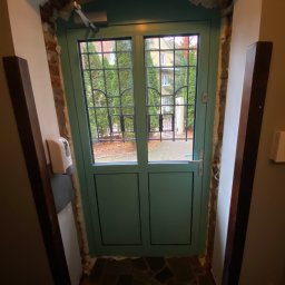 OMEGA Krzysztof Byckiewicz - Zielone aluminiowe drzwi wejściowe z kratką, widok z wnętrza budynku, z widocznymi śladami montażu i pianką uszczelniającą, automat samozamykający u góry