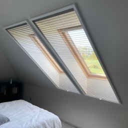 OMEGA Krzysztof Byckiewicz - Dwa okna dachowe z zamontowanymi plisami w sypialni na poddaszu, widok na zewnątrz przez okna.