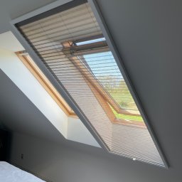 OMEGA Krzysztof Byckiewicz - Okno dachowe z plisowaną roletą w kolorze beżowym, częściowo zasłonięte, z widokiem na zewnątrz na zieleń drzew i niebieskie niebo, w tle fragment sypialni z białym łóżkiem.