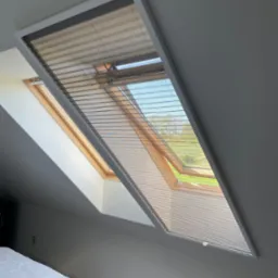 Okno dachowe z plisowaną roletą w kolorze beżowym, częściowo zasłonięte, z widokiem na zewnątrz na zieleń drzew i niebieskie niebo, w tle fragment sypialni z białym łóżkiem.