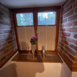 OMEGA Krzysztof Byckiewicz - Drewniane okno z plisowanymi roletami w rustykalnym wnętrzu z ceglaną ścianą, na parapecie dwie metalowe wazy, jedna z fioletowymi kwiatami.