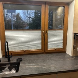 OMEGA Krzysztof Byckiewicz - Drewniane okno z plisami w kuchni nad zlewem z czarnym blatem.