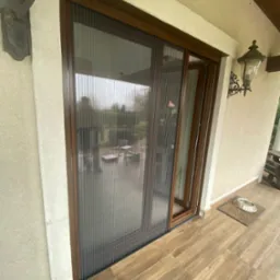 Zewnętrzna moskitiera plisowana w brązowej ramie, zamontowana na tarasie z drewnopodobną podłogą i widoczną lampą ścienną.