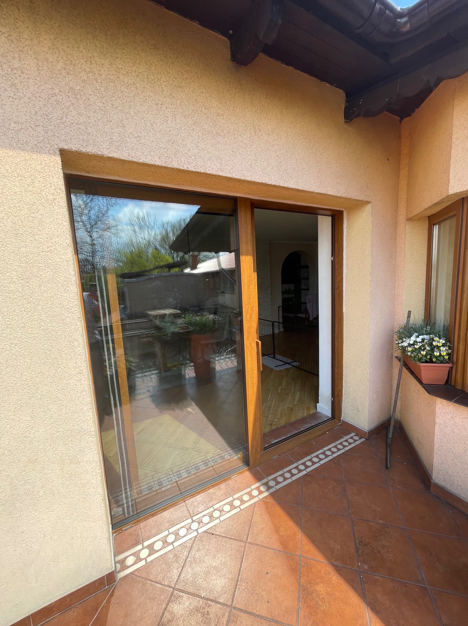 Przesuwne drzwi balkonowe z brązową ramą, widok z zewnątrz na taras z terakotową podłogą i roślinami w doniczce na parapecie.