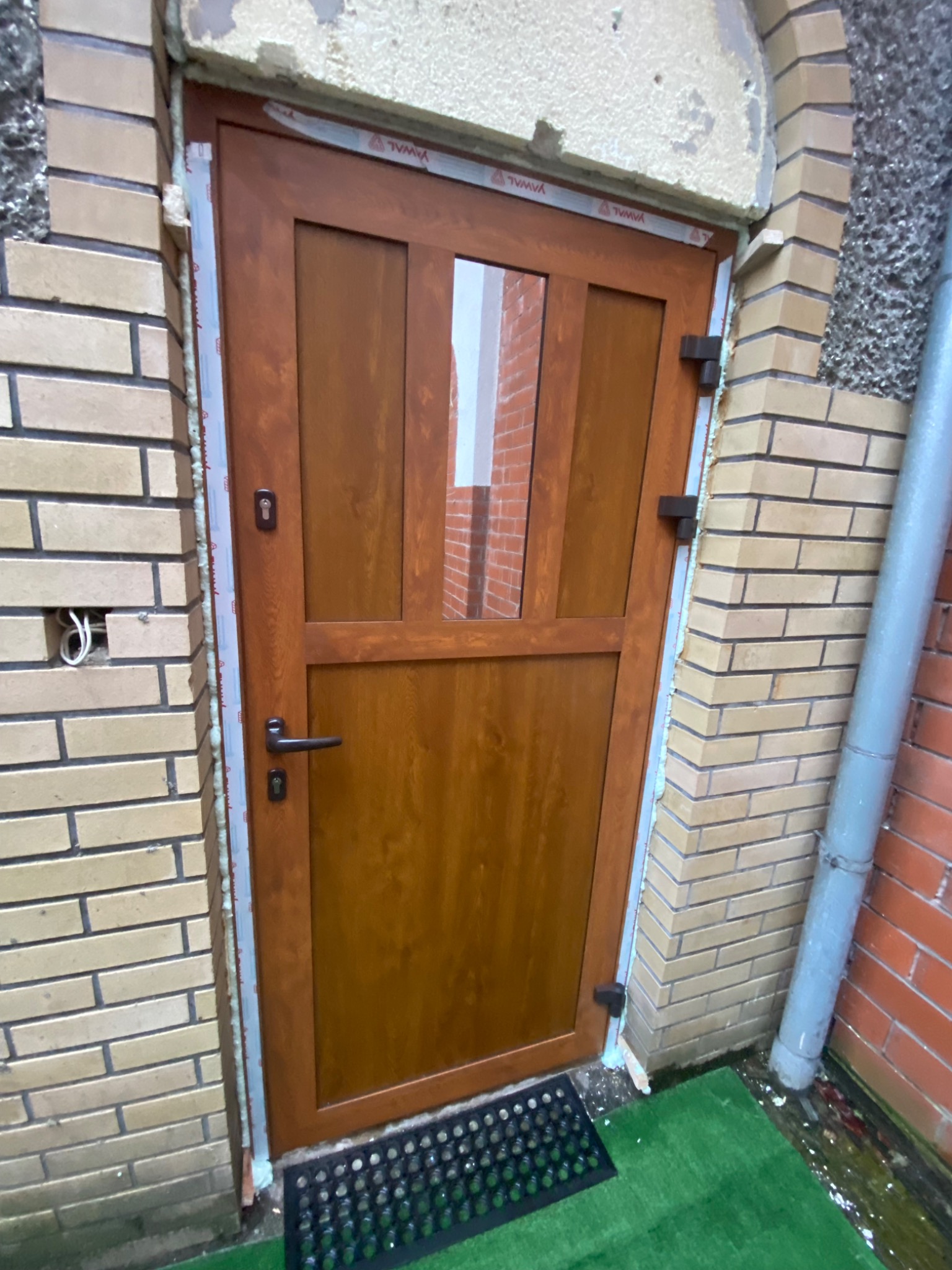 Brązowe drzwi wejściowe z szybą zamontowane w murze z cegły, widoczne ślady pianki montażowej, z zewnątrz widoczna instalacja elektryczna oraz rura spustowa.