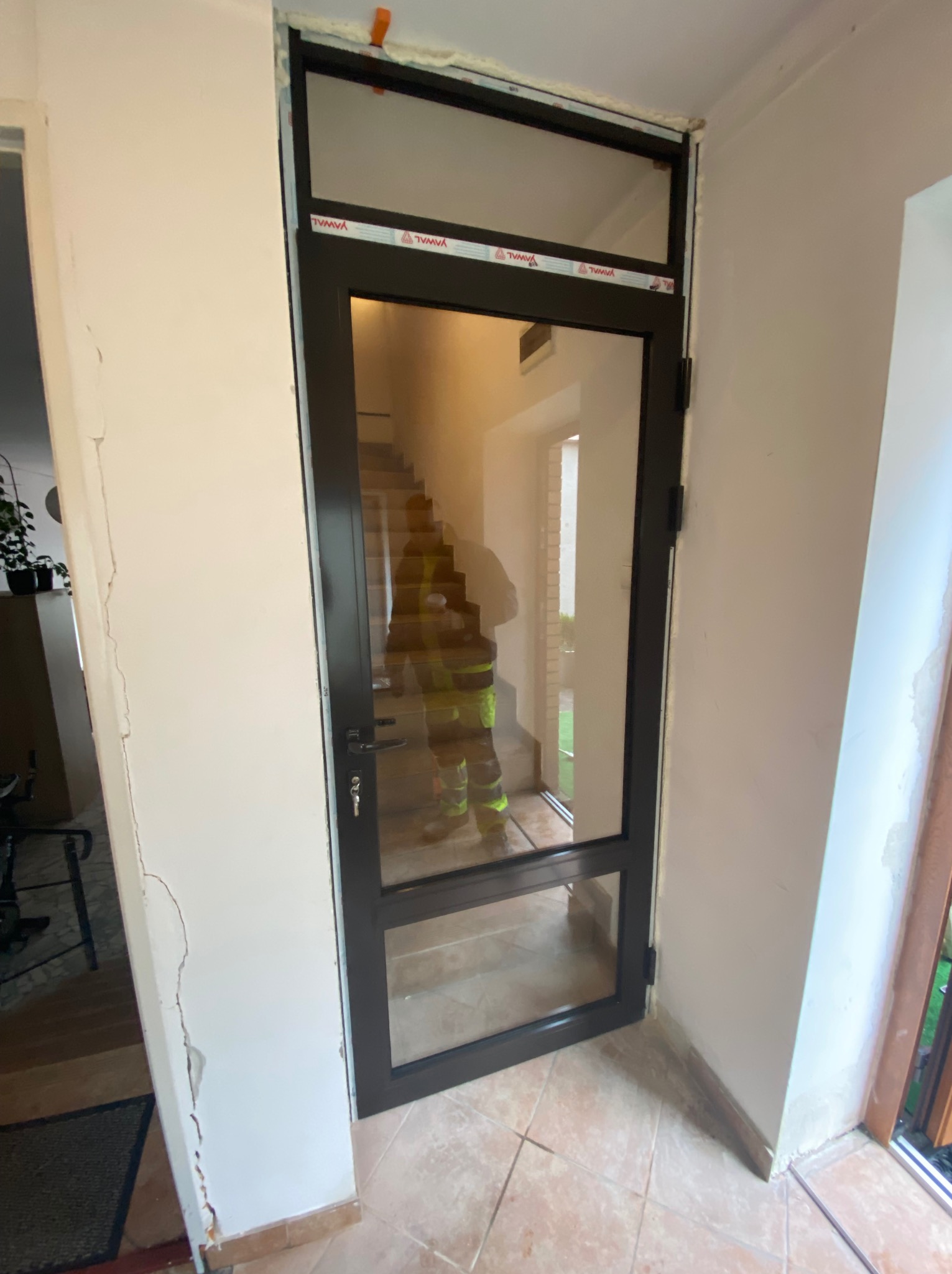 Czarna aluminiowa rama drzwiowa z dużym przeszkleniem wewnętrznym, widoczna osoba w odblaskowej kamizelce na schodach w tle, ślady pianki montażowej i uszkodzenia ściany.