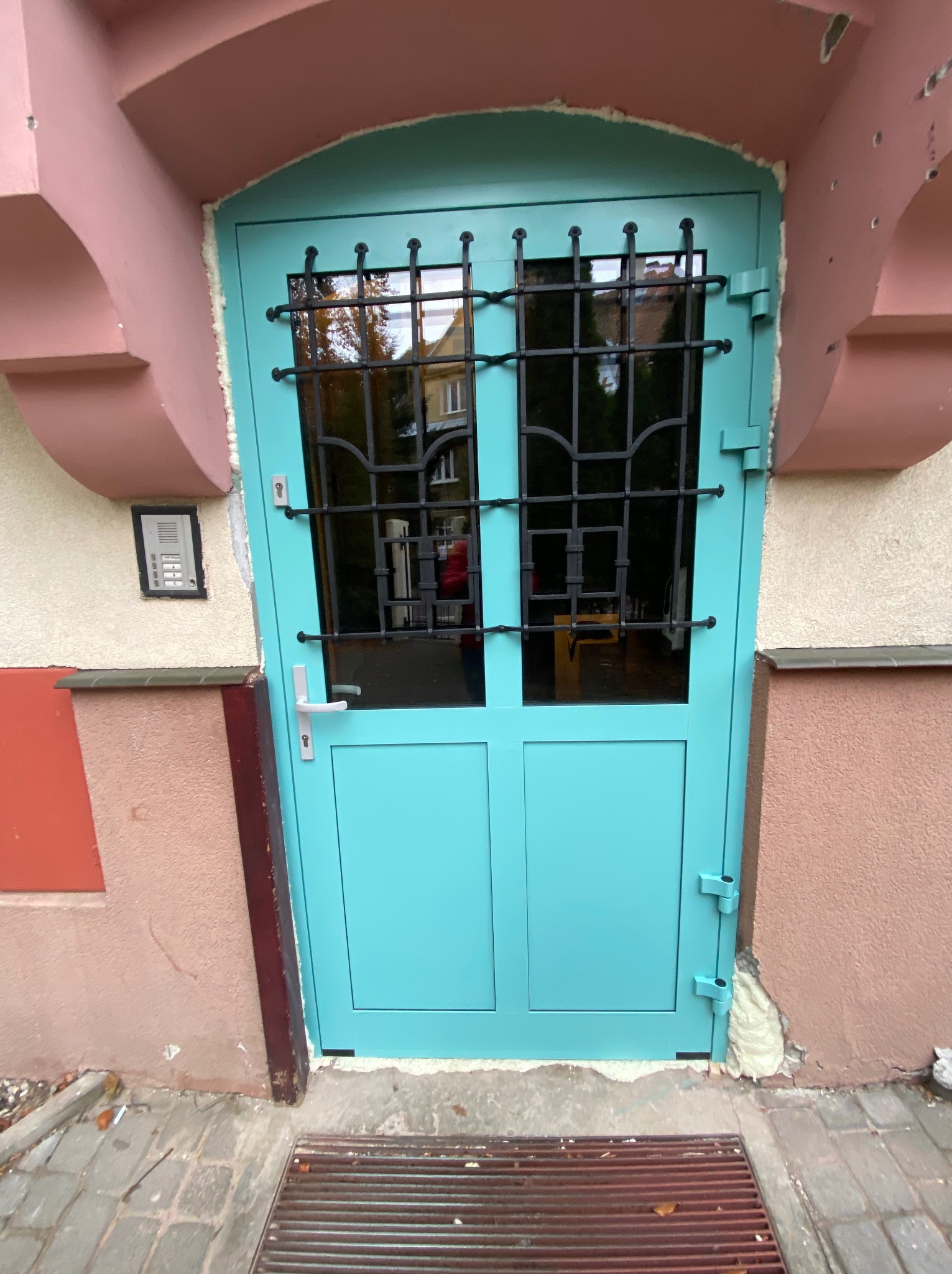 Turkusowe drzwi wejściowe z czarną kratą, osadzone w różowej fasadzie budynku, widoczne ślady pianki montażowej przy ościeżnicy.