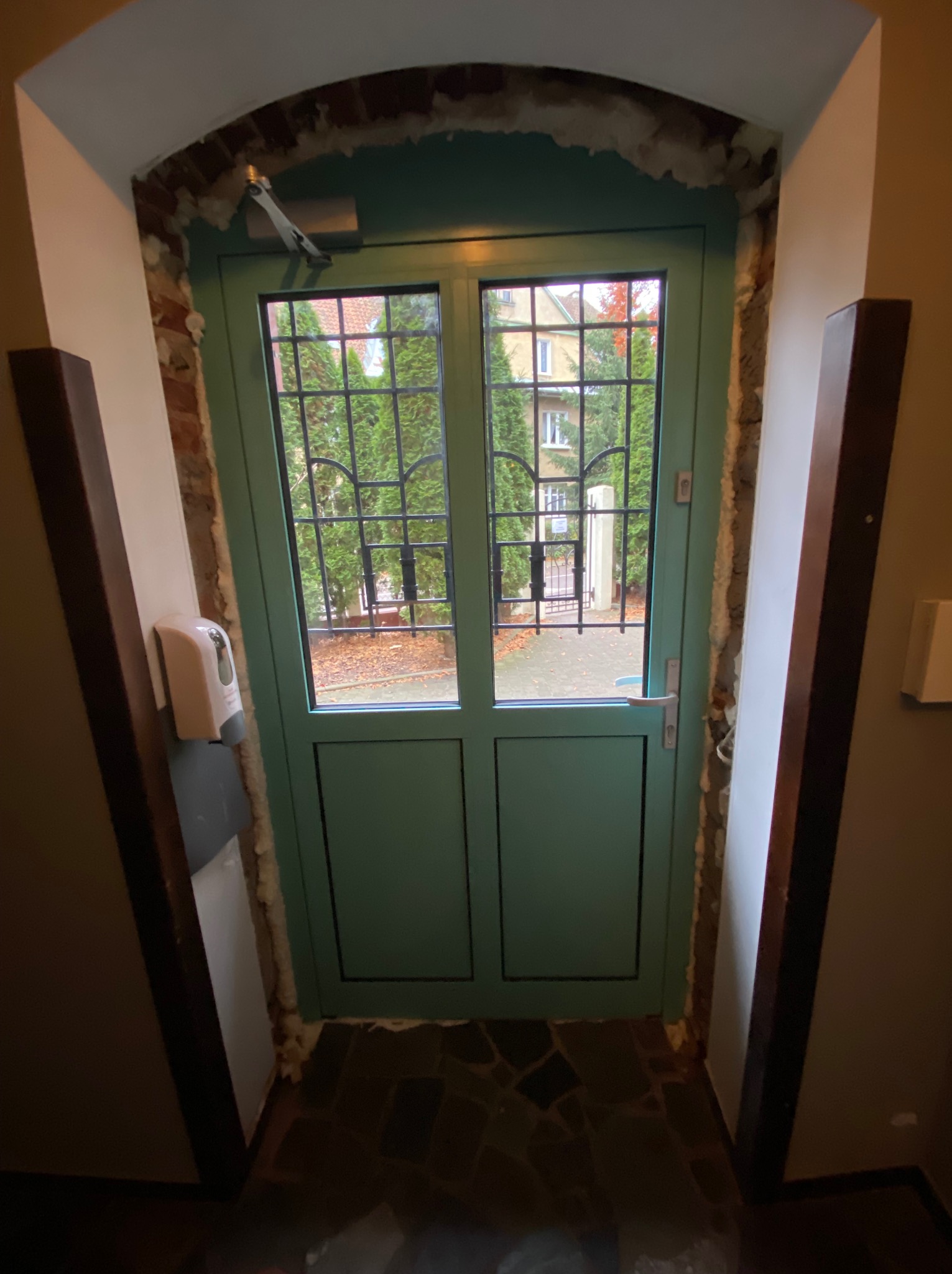 Zielone aluminiowe drzwi wejściowe z kratką, widok z wnętrza budynku, z widocznymi śladami montażu i pianką uszczelniającą, automat samozamykający u góry