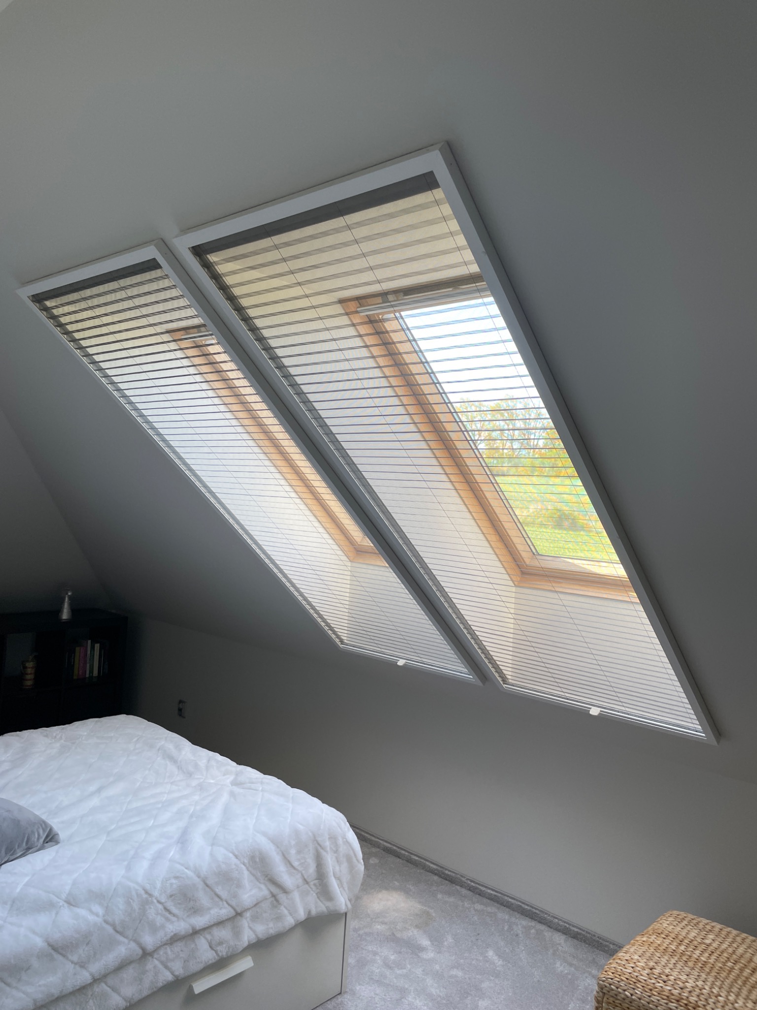 Dwa okna dachowe z zamontowanymi plisami w sypialni na poddaszu, widok na zewnątrz przez okna.