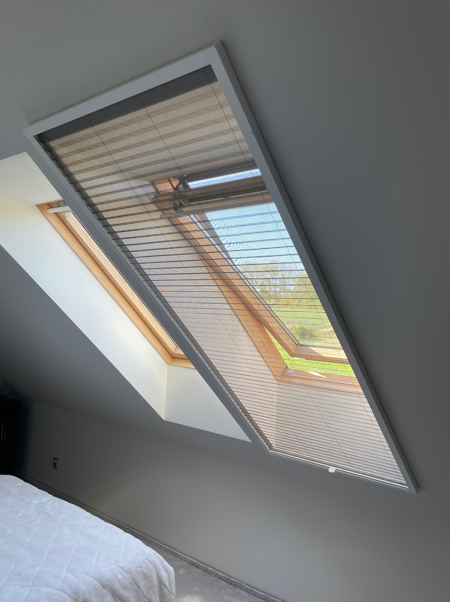 Okno dachowe z plisowaną roletą w kolorze beżowym, częściowo zasłonięte, z widokiem na zewnątrz na zieleń drzew i niebieskie niebo, w tle fragment sypialni z białym łóżkiem.