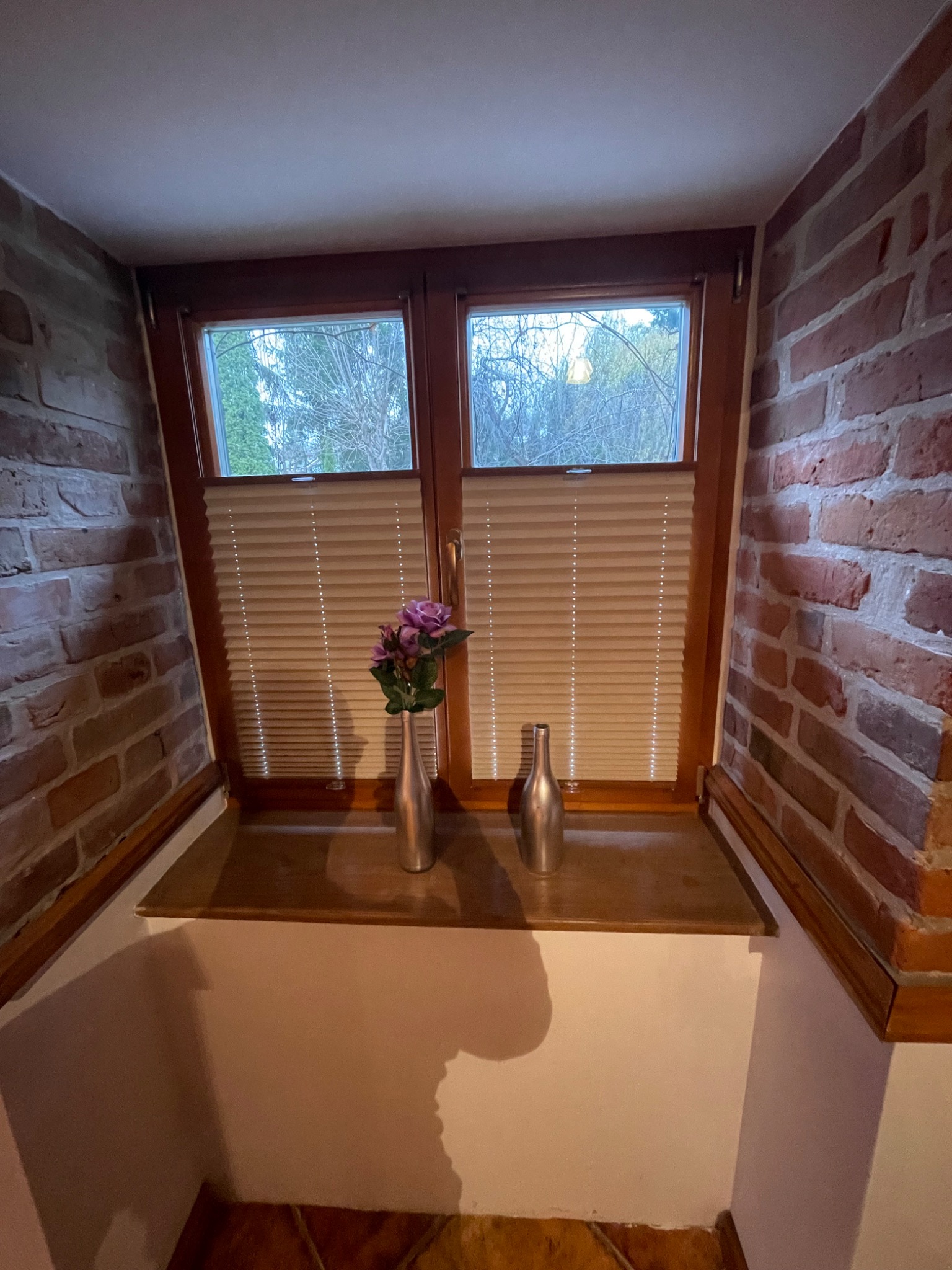 Drewniane okno z plisowanymi roletami w rustykalnym wnętrzu z ceglaną ścianą, na parapecie dwie metalowe wazy, jedna z fioletowymi kwiatami.