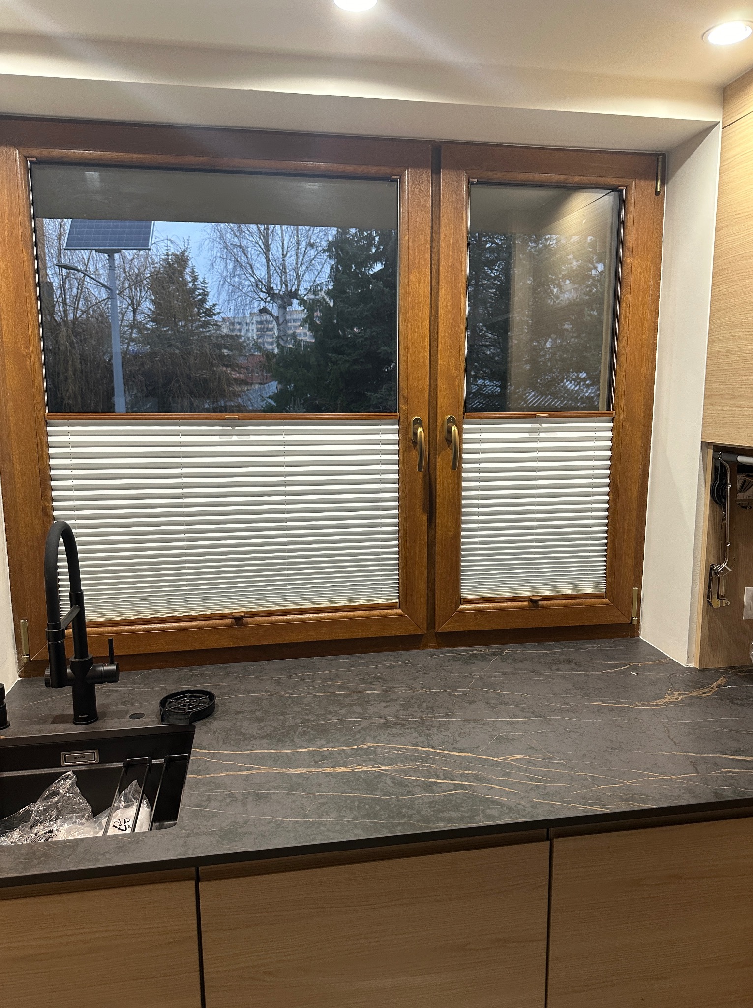 Drewniane okno z plisami w kuchni nad zlewem z czarnym blatem.