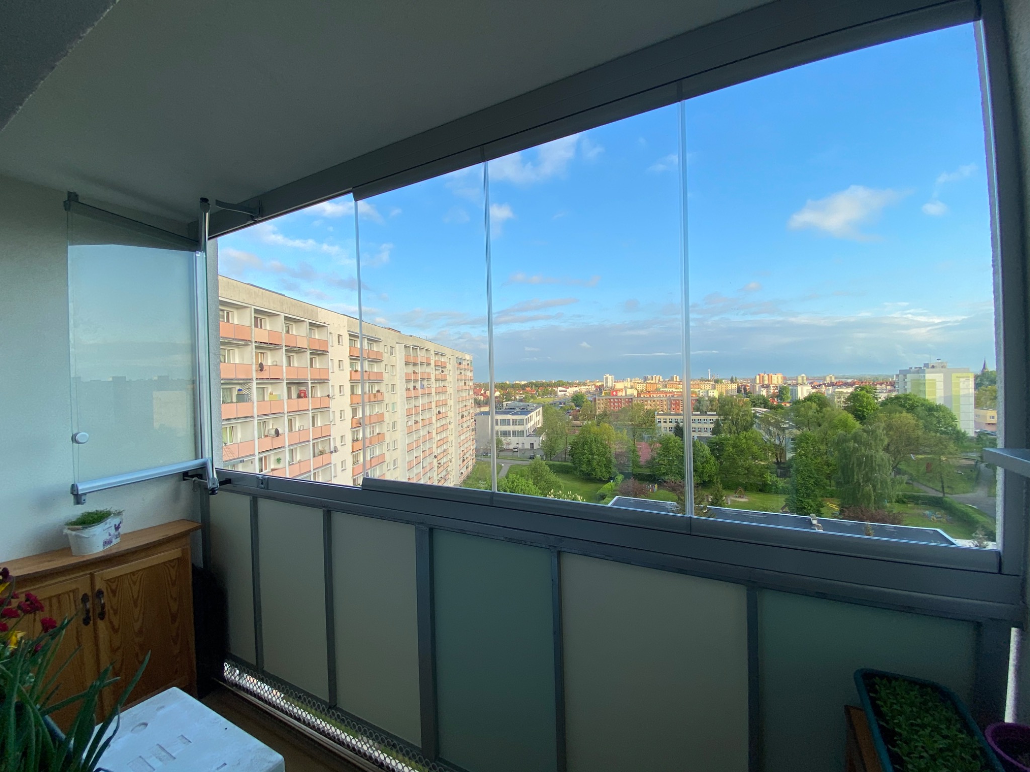 Zabudowany balkon z przesuwnymi szklanymi panelami, widok na osiedle mieszkaniowe w pogodny dzień.