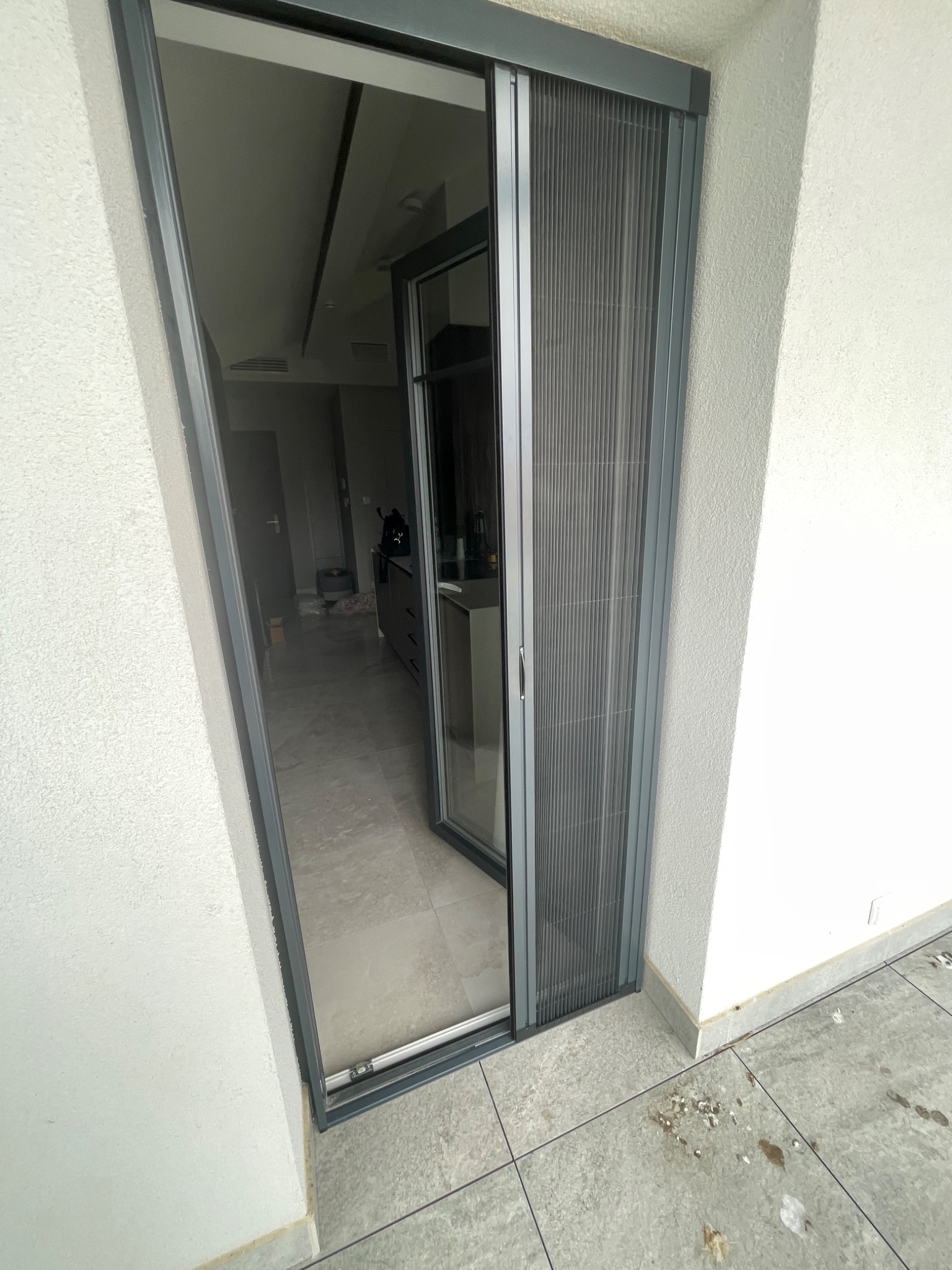 Szara przesuwna moskitiera plisowana w otworze drzwi balkonowych, widok z zewnątrz, zarys wnętrza mieszkania w tle.