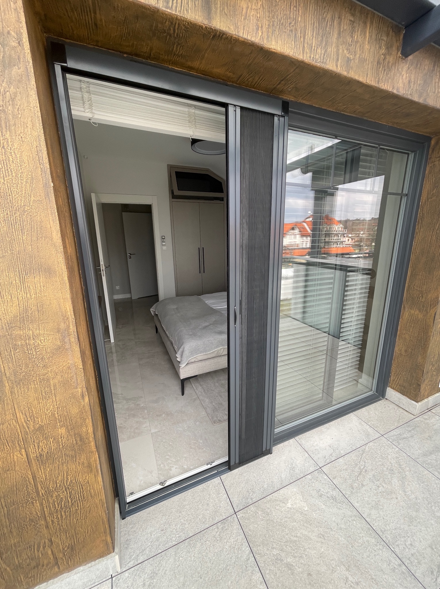 Przesuwne okno balkonowe z szarą ramą i plisowaną moskitierą, widok na sypialnię z łóżkiem i zabudowaną szafą, za oknem widoczny budynek z czerwonym dachem.