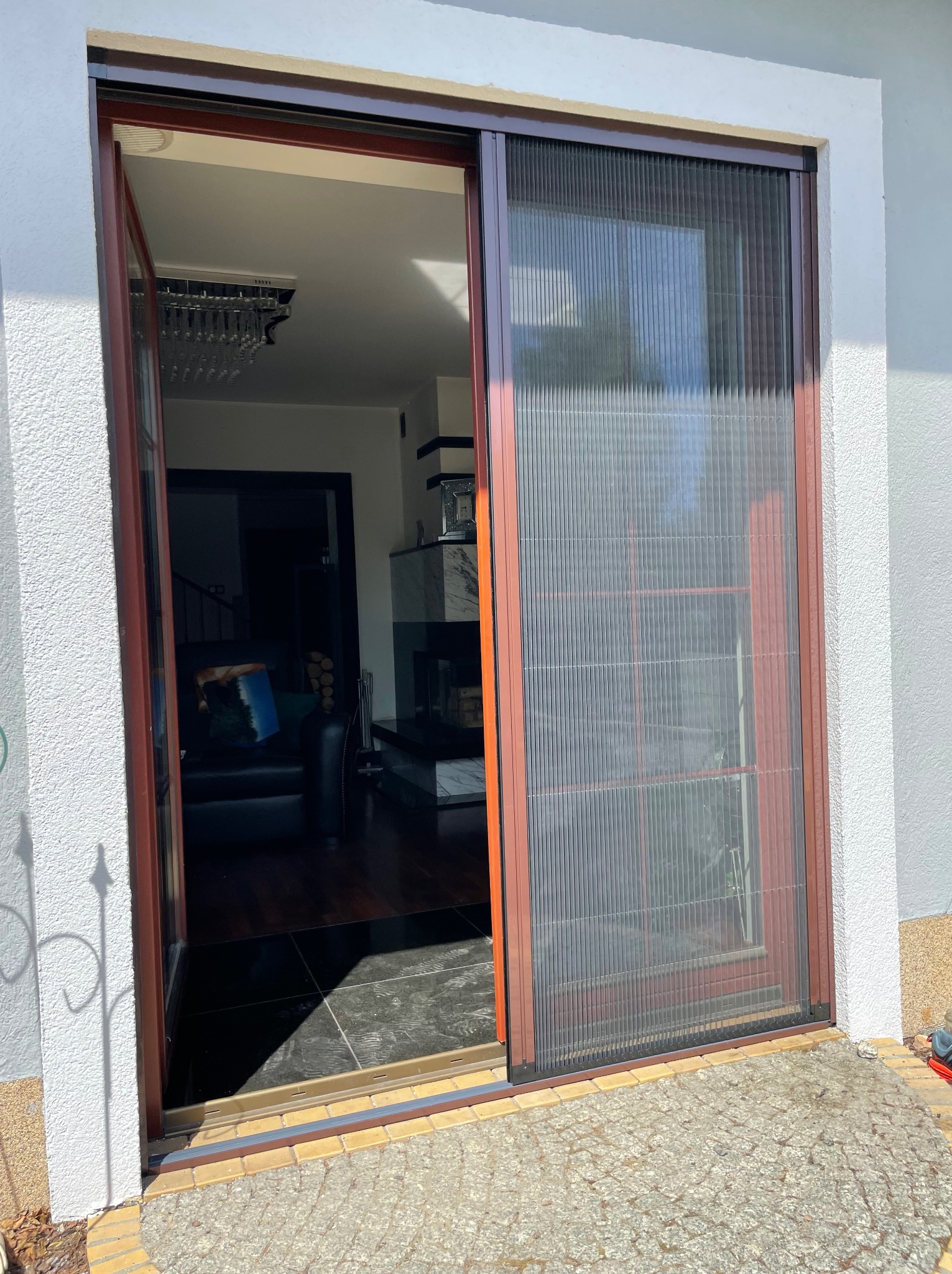 Rozsunięta moskitiera plisowana w brązowej ramie na tle białej ściany domu, widok na wnętrze salonu z kominkiem i schodami.